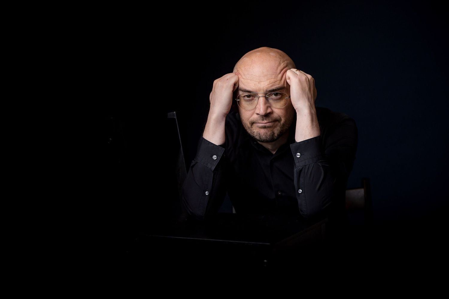 Stressed bald man with glasses holding his head in his hands at a dark desk, looking frustrated.