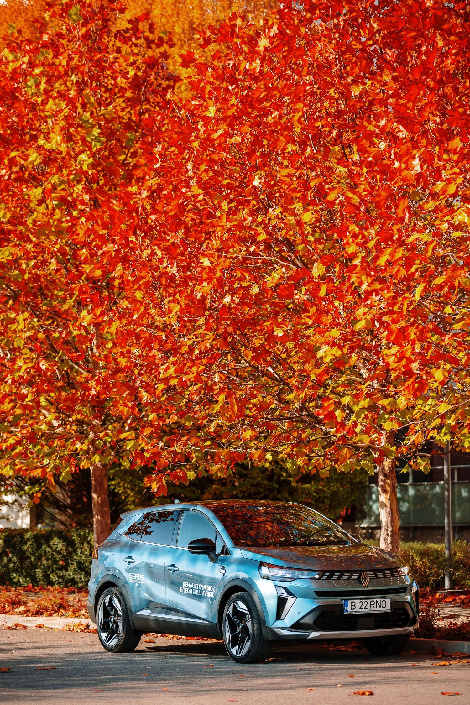 Blue electric SUV parked beneath vibrant red and orange autumn trees on a sunny fall day.