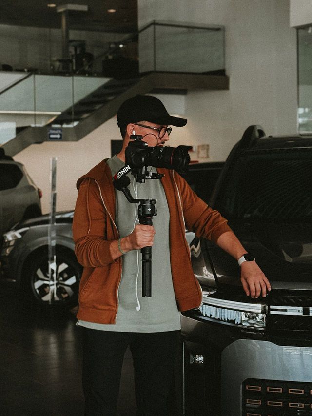 Bogdan Grigore - videographer in a brown hoodie and black cap films with a camera gimbal stabilizer in a modern car showroom.