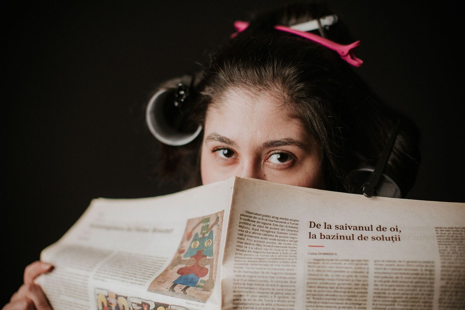 Young girl with pink hair clips reads a newspaper, covering her face, with curious eyes peeking over the top.