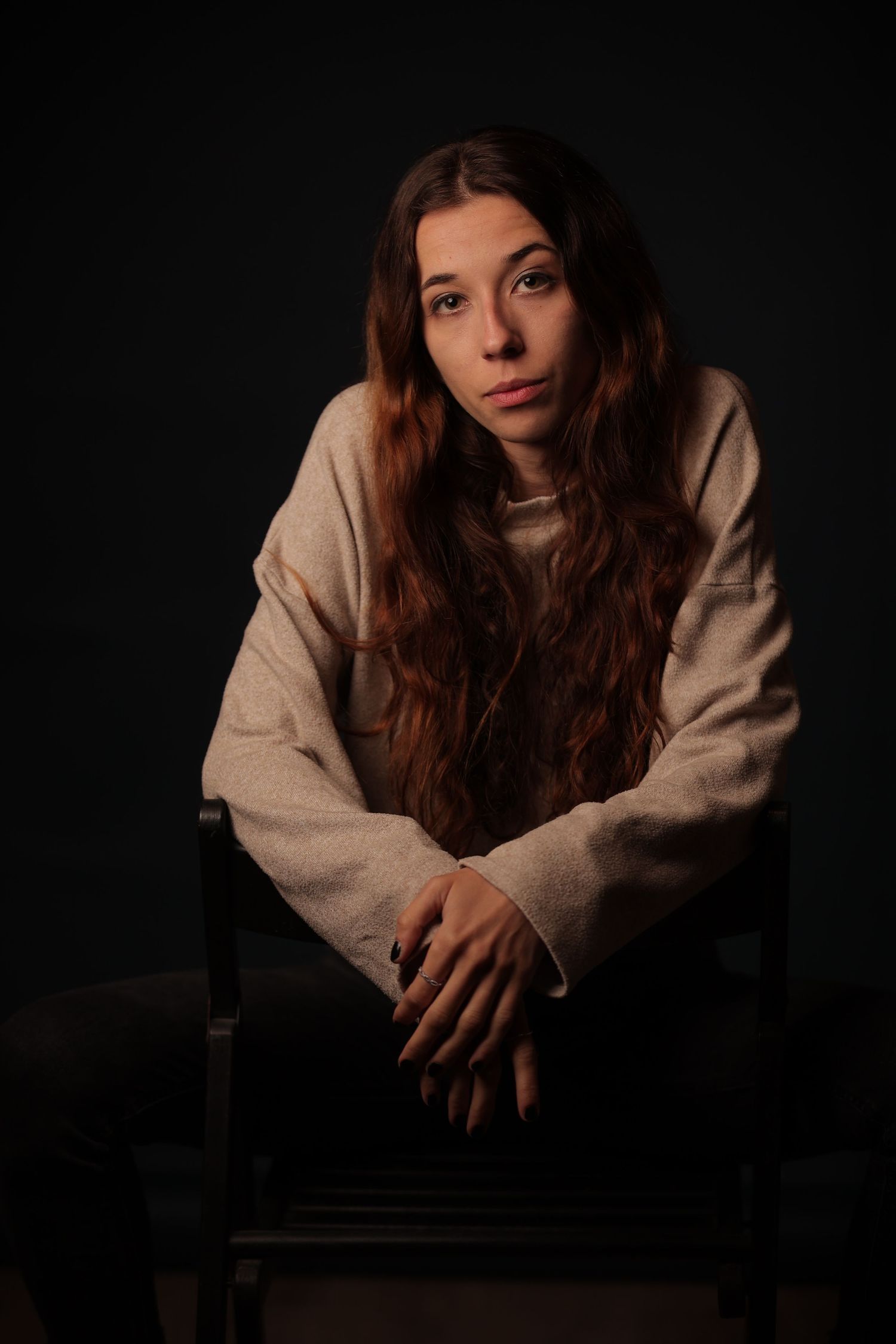 Young woman with long wavy auburn hair sits on a chair wearing a beige sweatshirt against a dark studio background.
