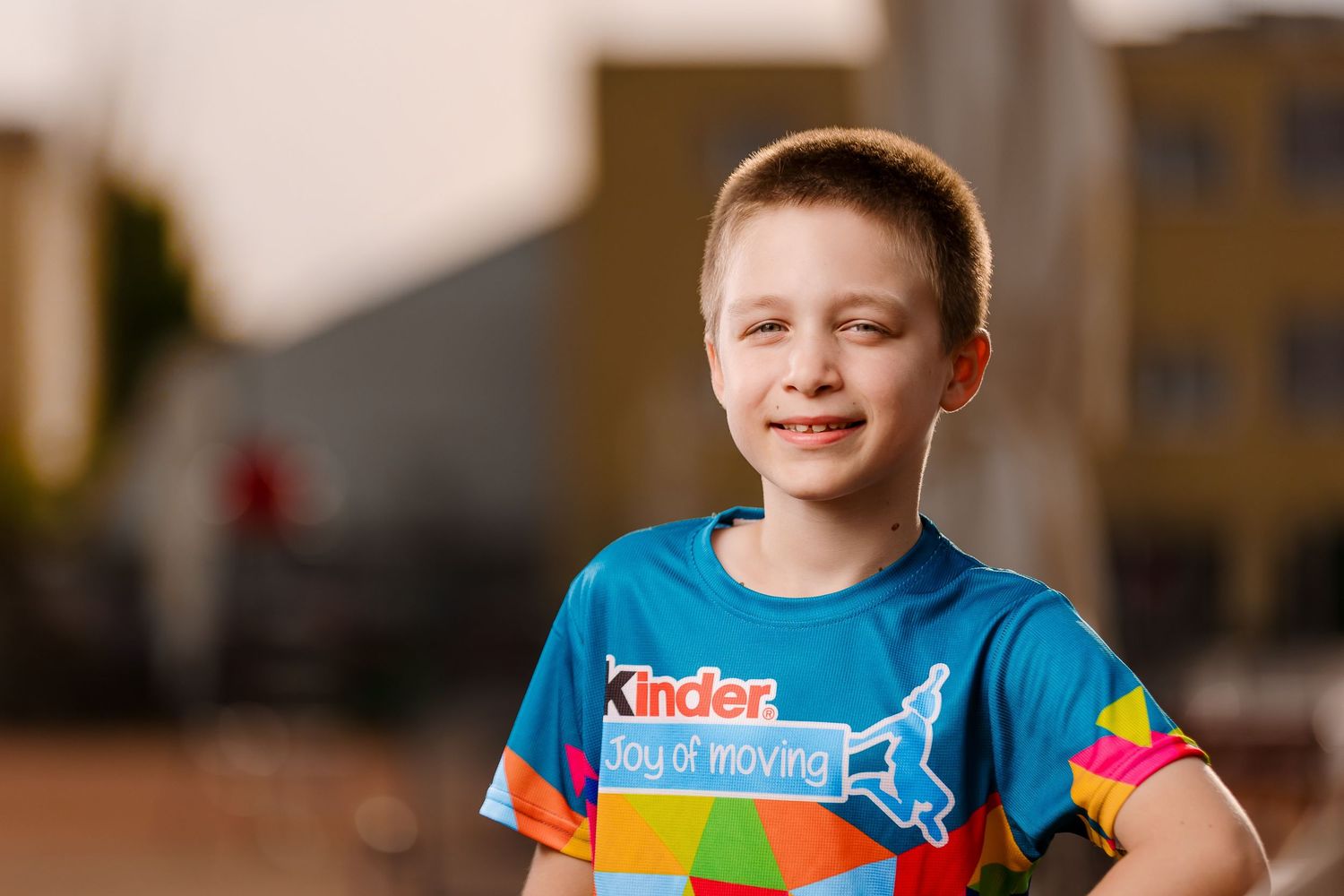 Young boy smiling in a blue Kinder Joy of Moving t-shirt outdoors during a sports event.