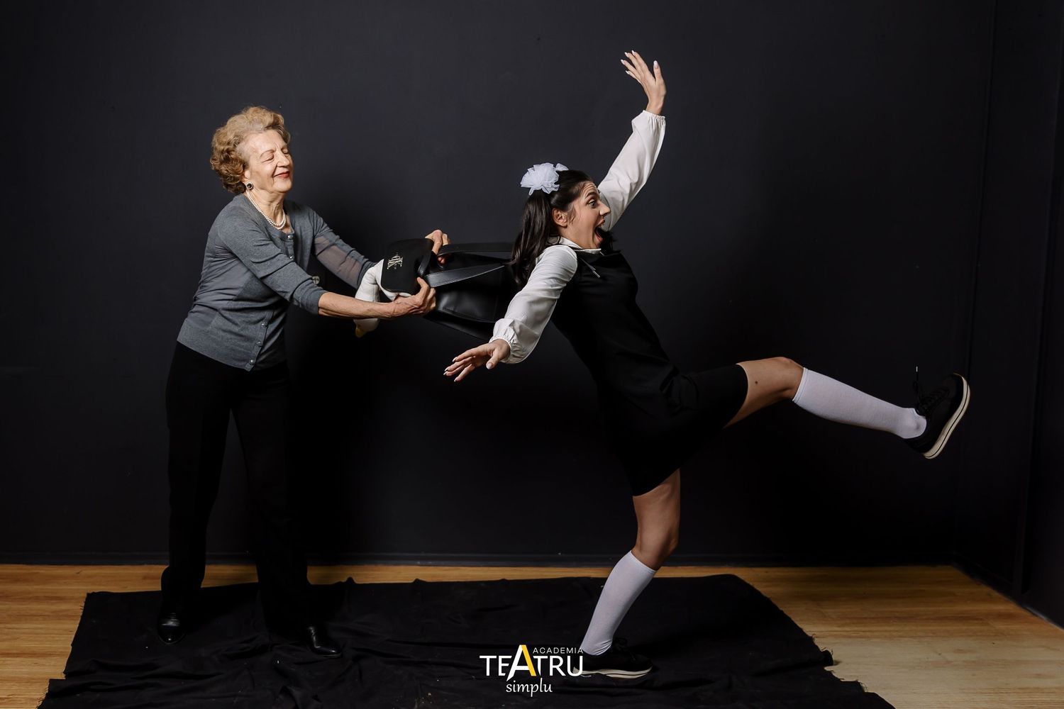 Two female theater performers interact dynamically, one balancing on one leg with arm raised on a black stage.