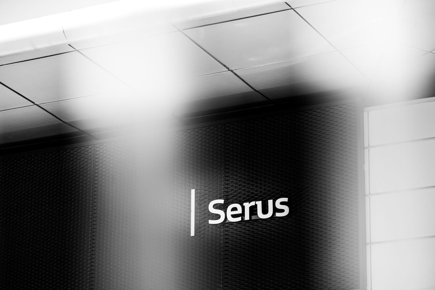 Black and white photo of a modern office sign reading Serus on a dark textured wall.