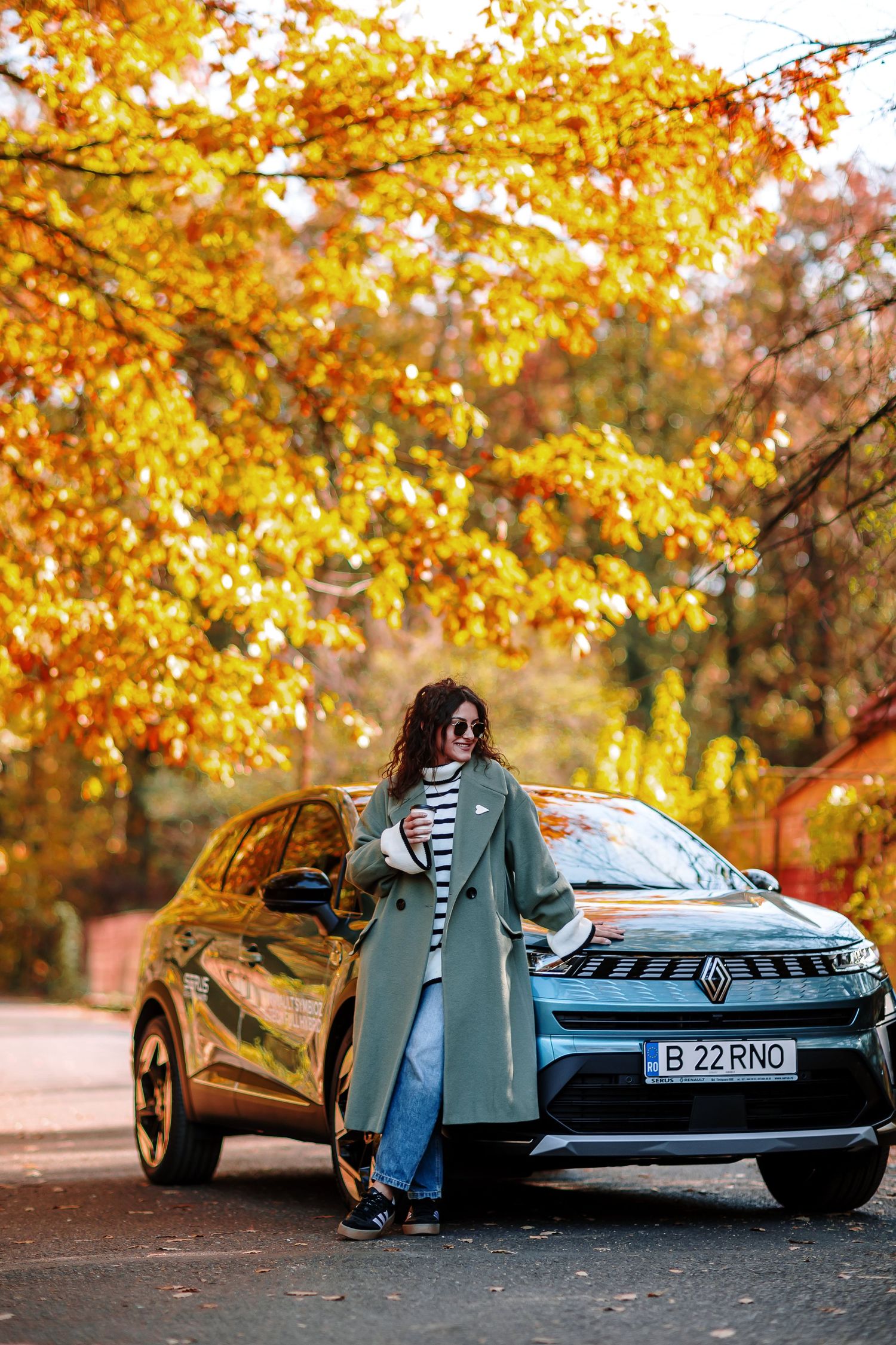 Woman in green coat leaning on Renault car amid vibrant autumn foliage on a scenic road.