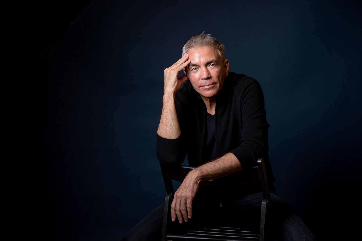 Middle-aged man with gray hair in black outfit poses thoughtfully against dark blue studio background.
