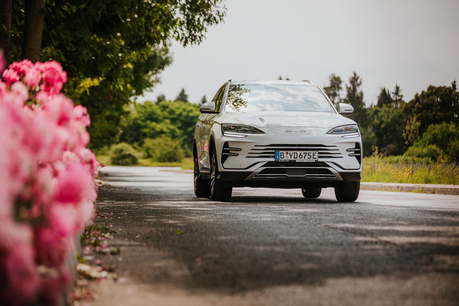 Modern electric SUV driving on a road surrounded by lush greenery and pink flowering bushes on a sunny day.
