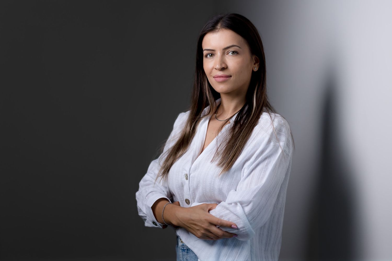 A confident woman in a white blouse stands with arms crossed against a dark studio background.