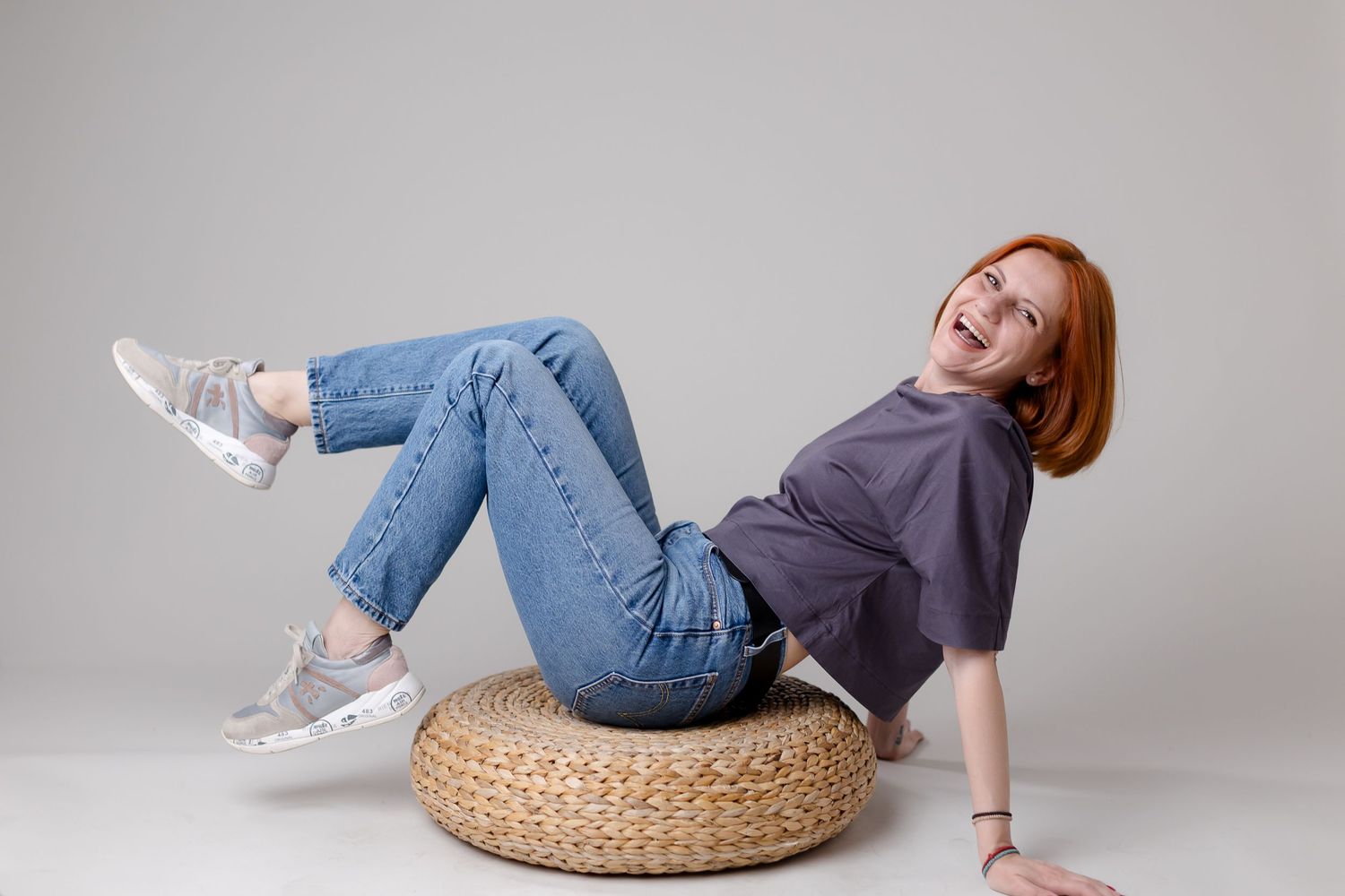 Laughing redhead woman in jeans and gray tee sits on woven pouf against white background.