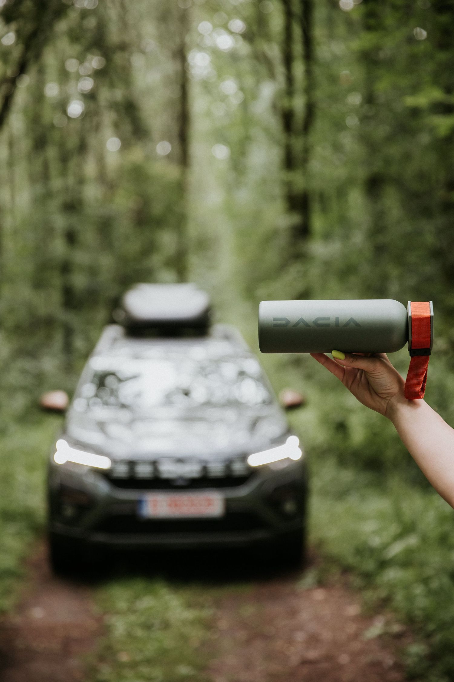 Hand holding a Dacia branded green water bottle in a forest with a Dacia SUV on a dirt trail in the background.