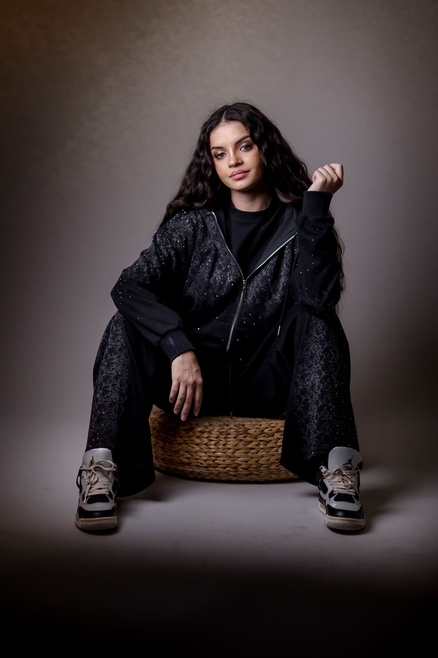 Young woman in black tracksuit and sneakers sitting on wicker basket, posing confidently against gray studio background.