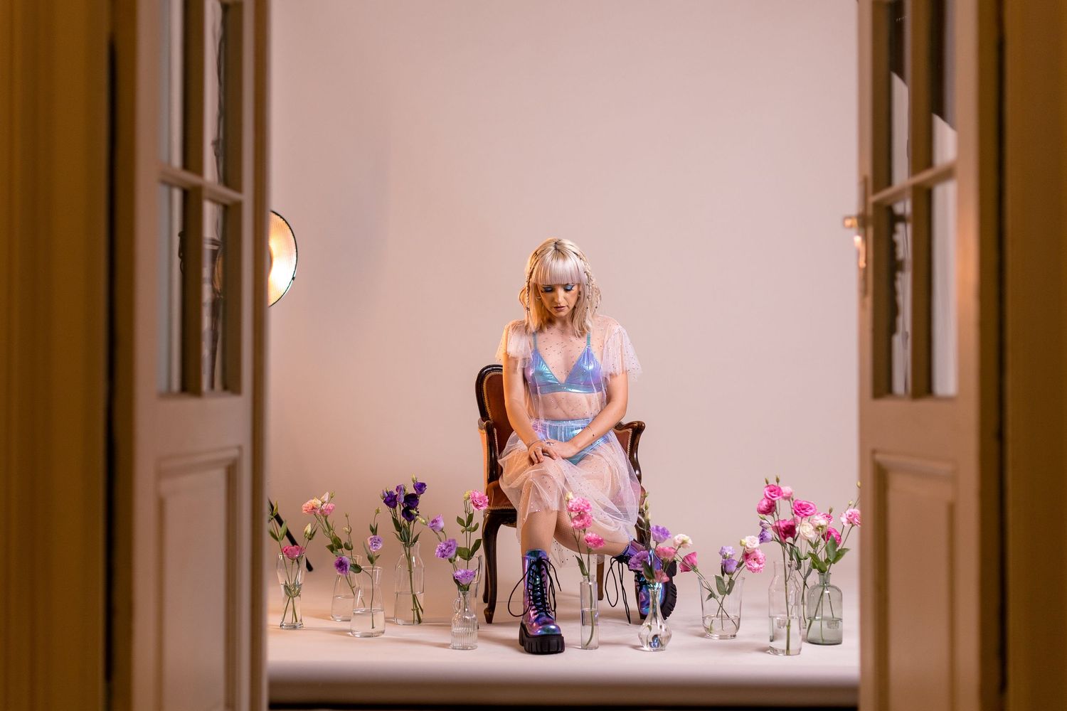 Blonde woman sitting on chair surrounded by colorful flowers in glass vases, wearing blue lingerie top and pink skirt.