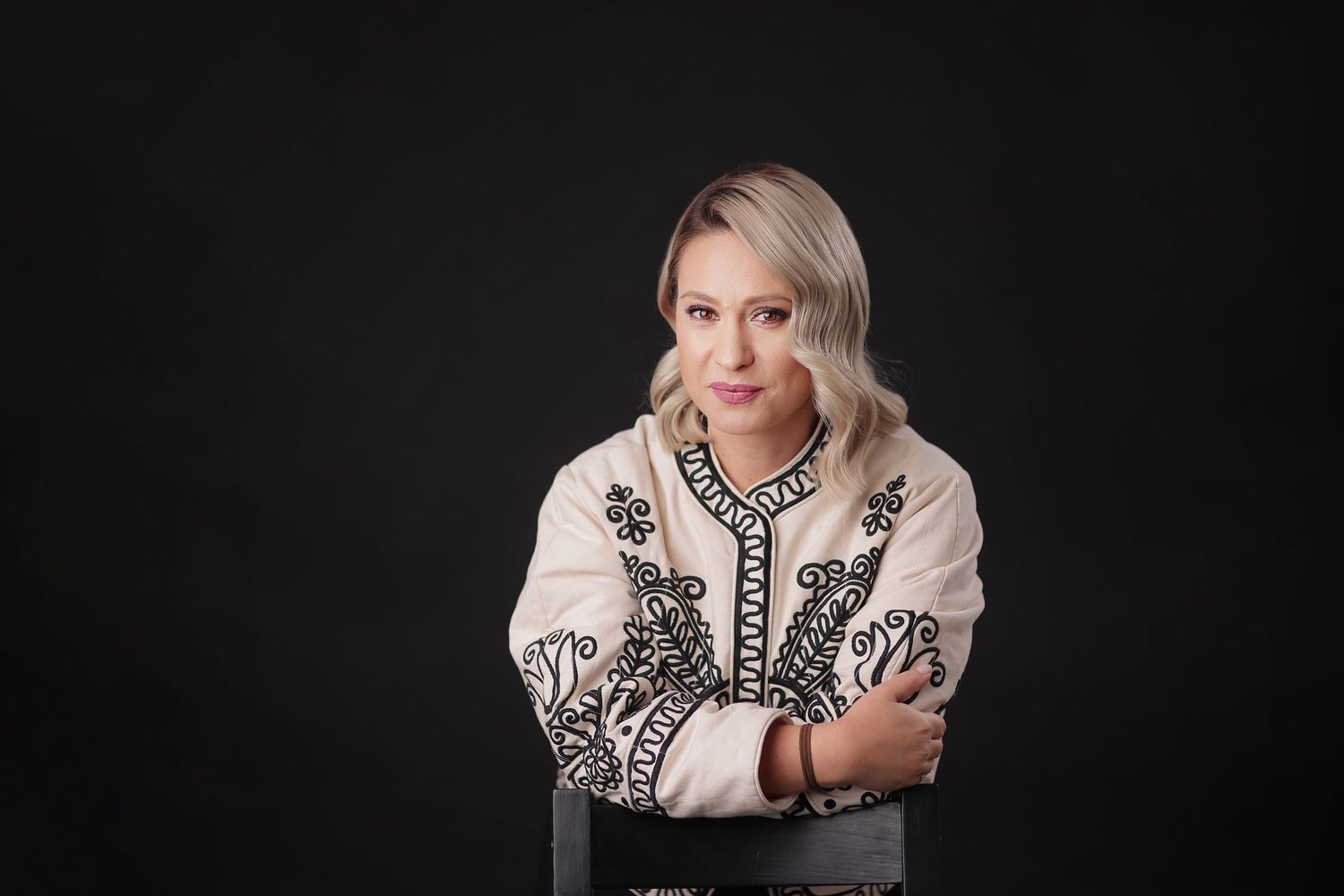 Blonde woman in embroidered white jacket sits with arms crossed on dark background.