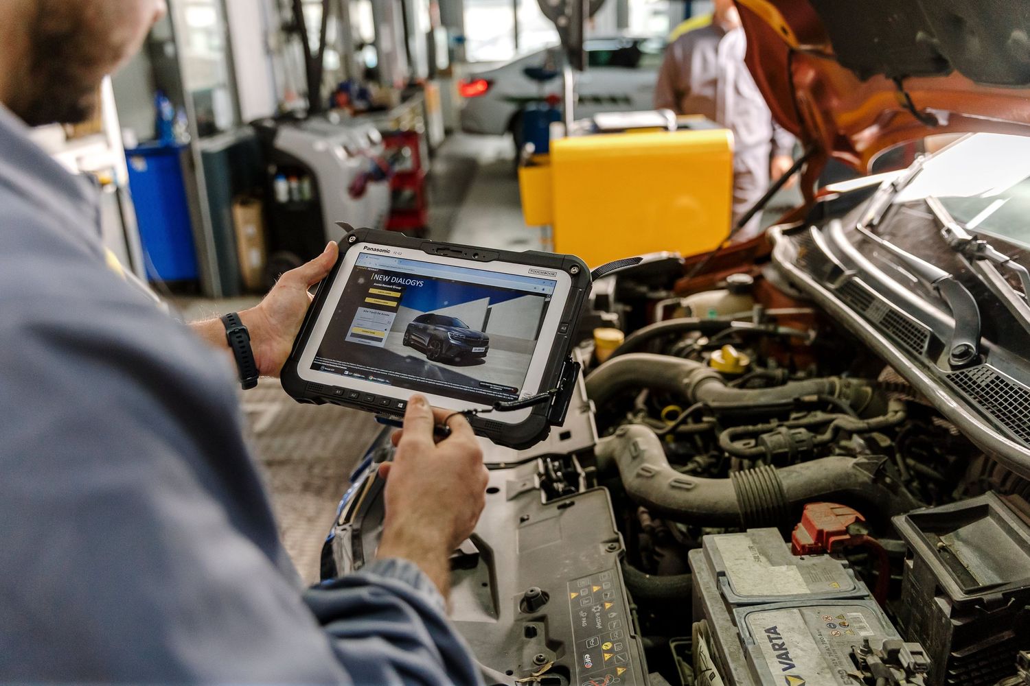 Mechanic using a rugged tablet to diagnose a car engine in an auto repair shop.