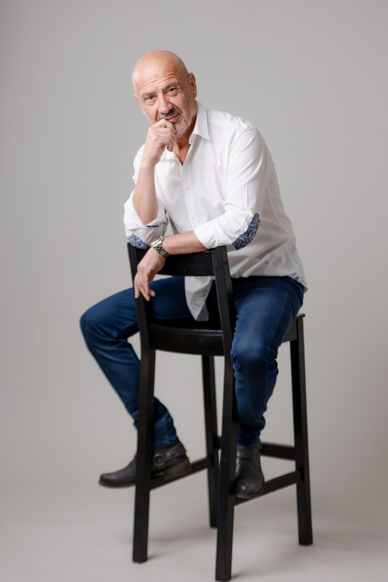Bald middle-aged man in white shirt and blue jeans sitting on a black bar stool with hand on chin.