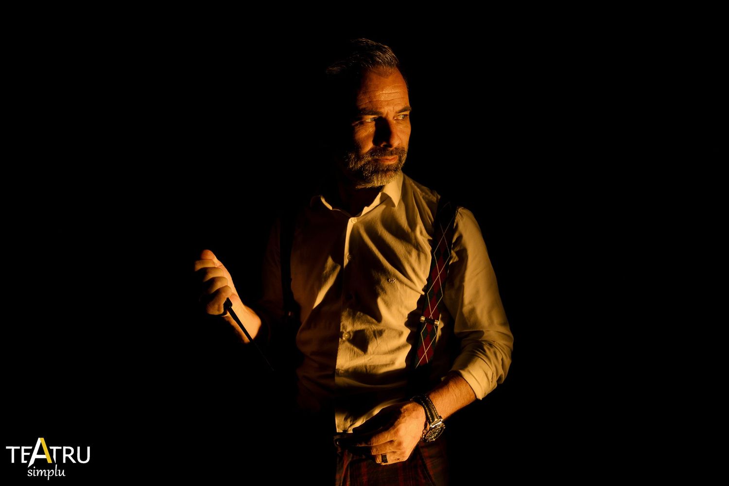 A dramatic male theater actor poses in dim amber light wearing a vest and white shirt on a dark stage.