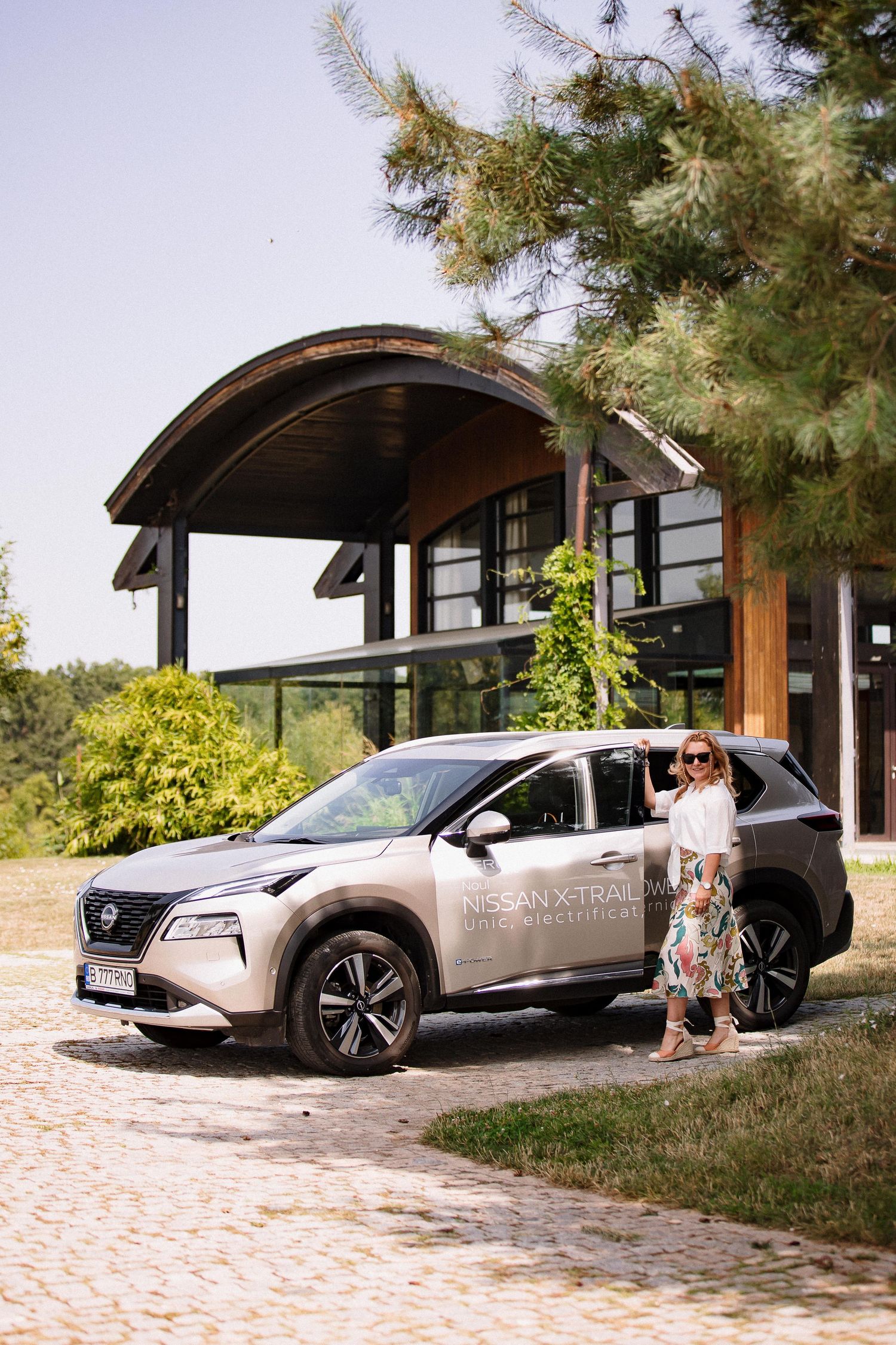 Woman standing beside silver Nissan X-Trail SUV in front of modern wooden building on sunny day.