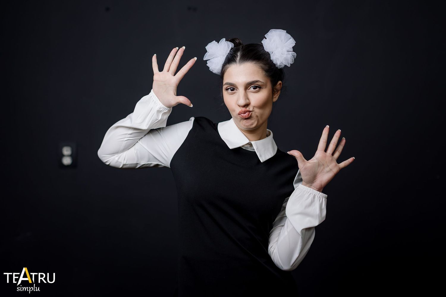 A performer in black dress with white collar and pom-pom headband poses dramatically against dark background.