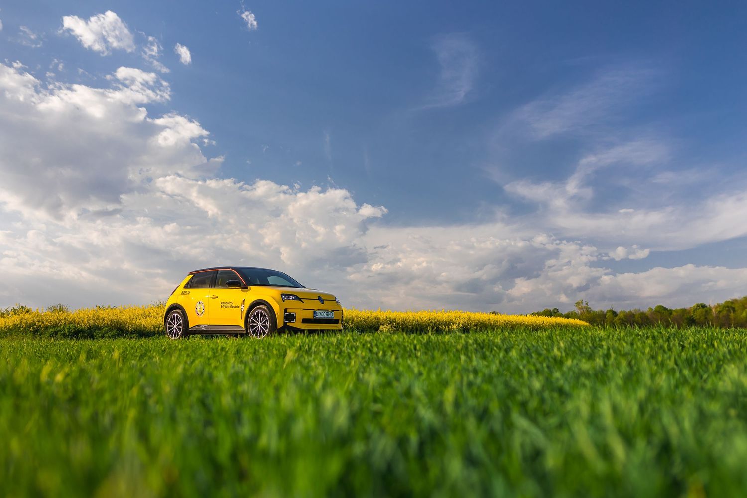 Yellow compact car parked in a lush green field under a bright blue sky with fluffy white clouds on a sunny day.