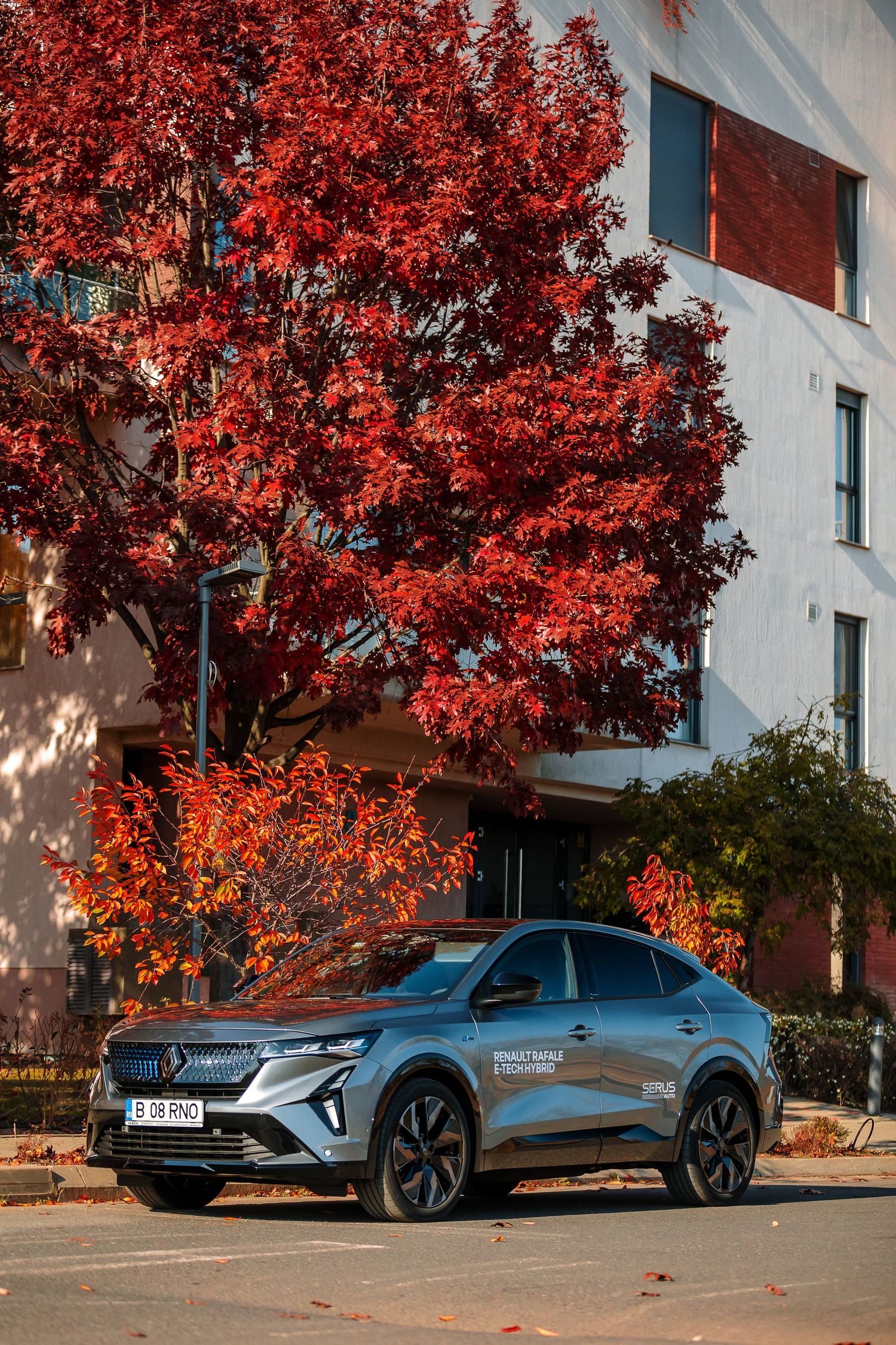 Silver Renault SUV parked beside vibrant red autumn trees near a modern white building on a sunny day.