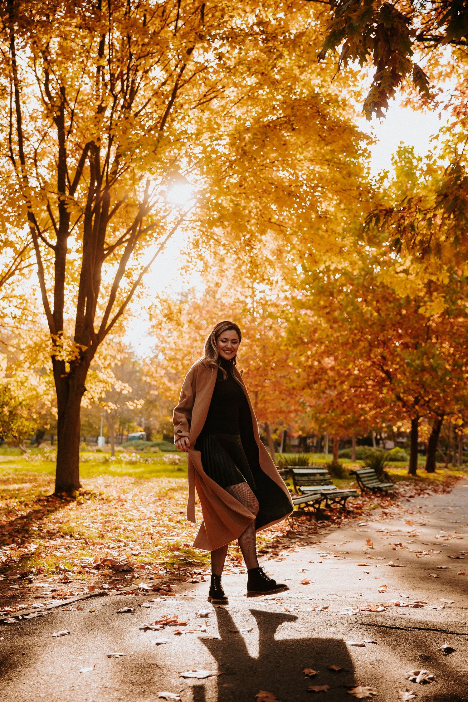 Woman in autumn park wearing brown coat and skirt, smiling on wet path surrounded by golden fall foliage.
