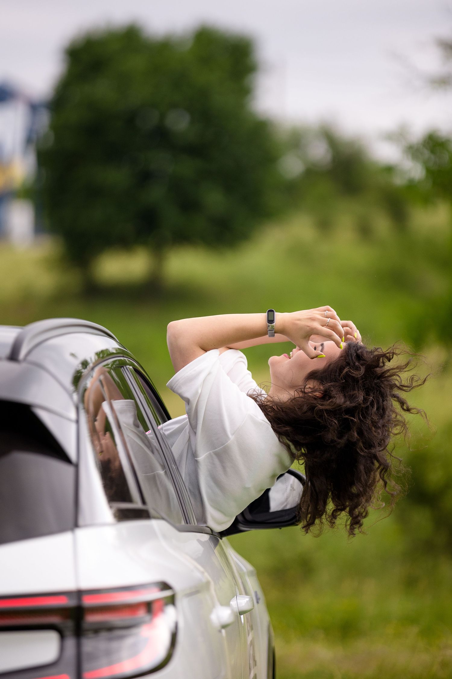 Young woman with curly hair leaning out of a white car window, enjoying a scenic green countryside drive.