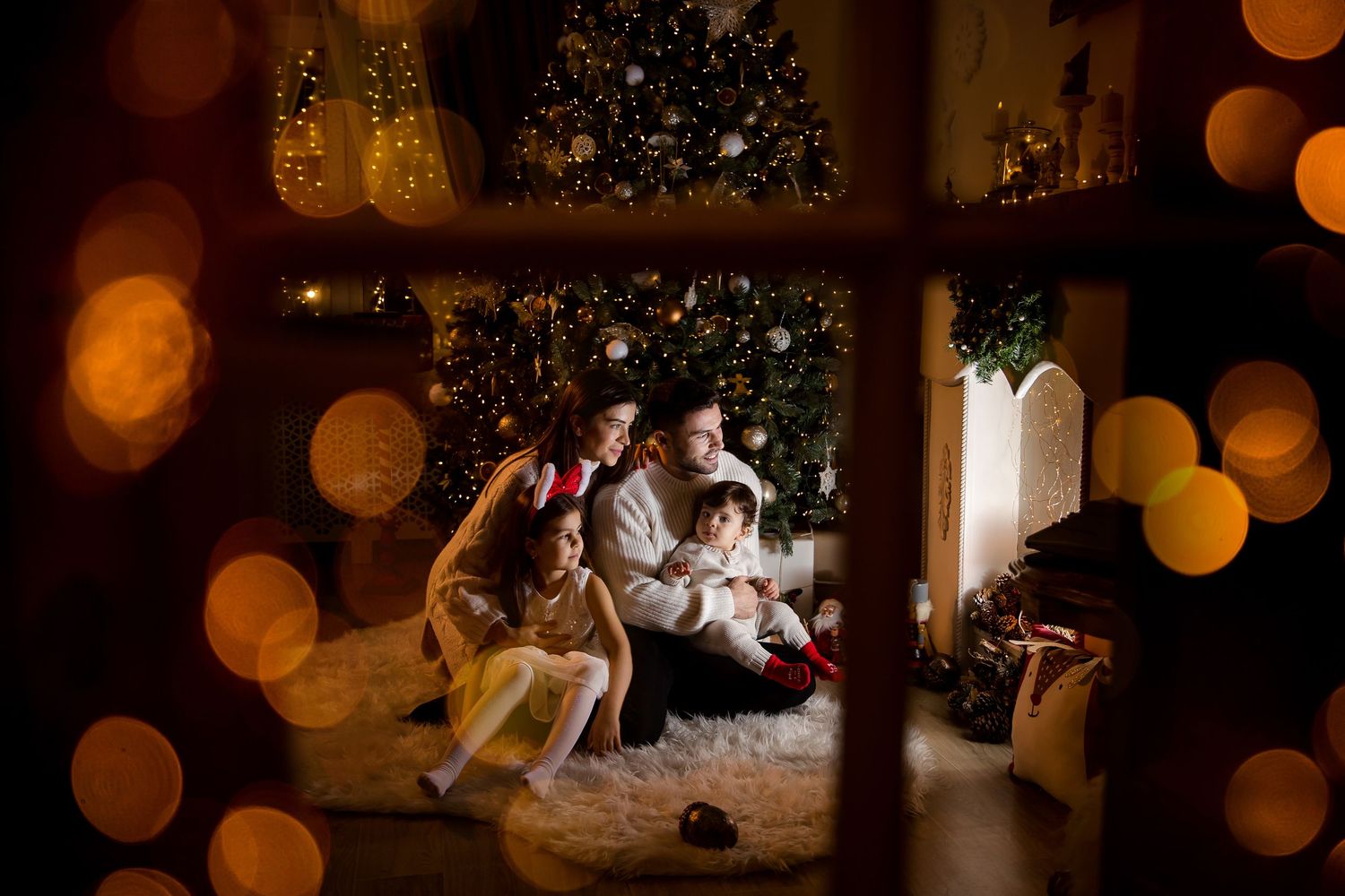 Family gathered by a decorated Christmas tree, viewed through a window with warm bokeh lights.