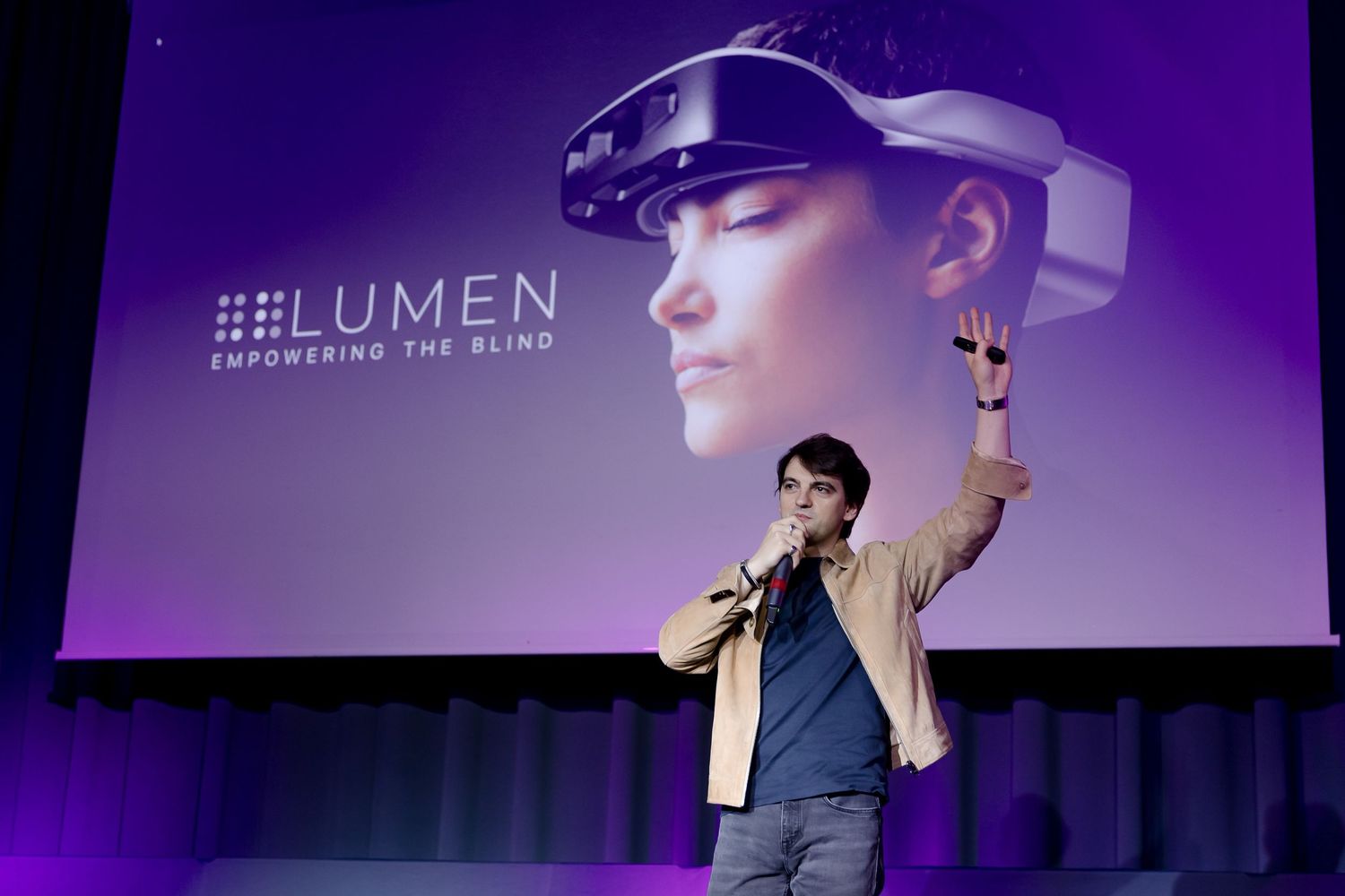 A presenter holds up a VR headset on stage at a Lumen Empowering the Blind tech conference presentation.