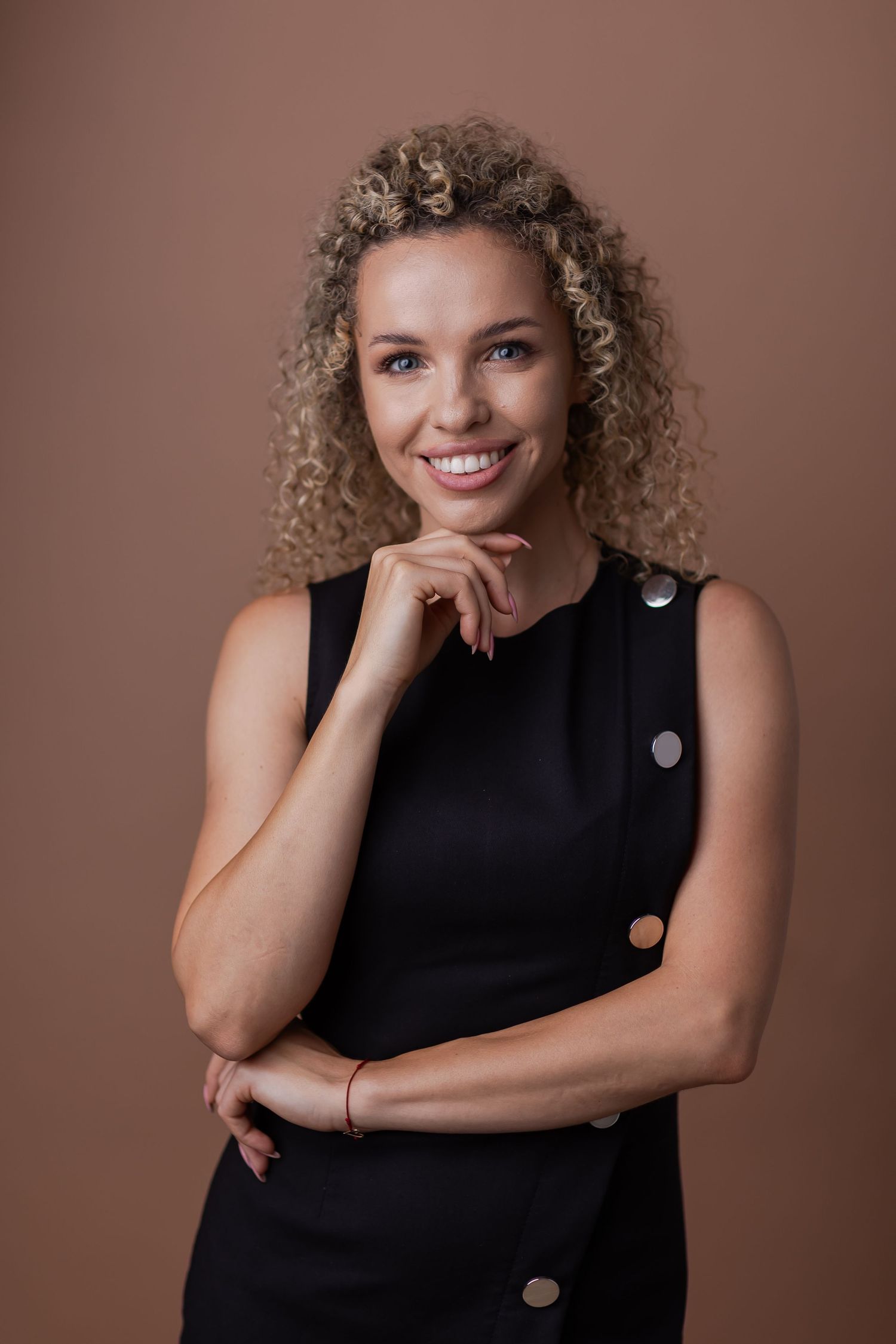 Smiling woman with curly blonde hair in black sleeveless dress, hand on chin, against brown background.