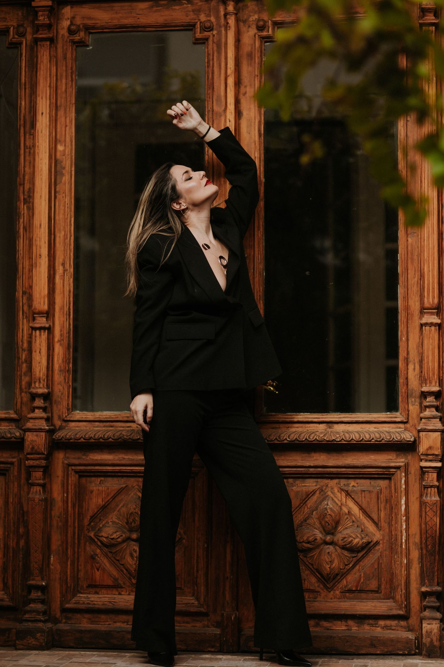 Woman in black suit posing confidently against ornate wooden doors, one arm raised, long hair flowing.