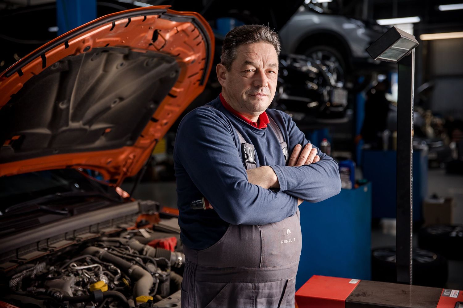 Confident auto mechanic with crossed arms standing in front of an orange car with open hood in repair shop.