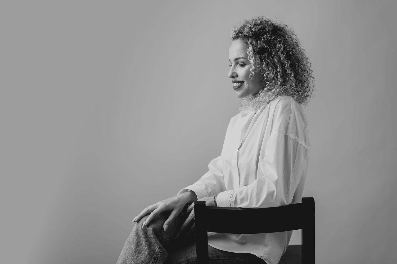 Black and white portrait of a curly-haired woman sitting on a chair, smiling softly in a white blouse.
