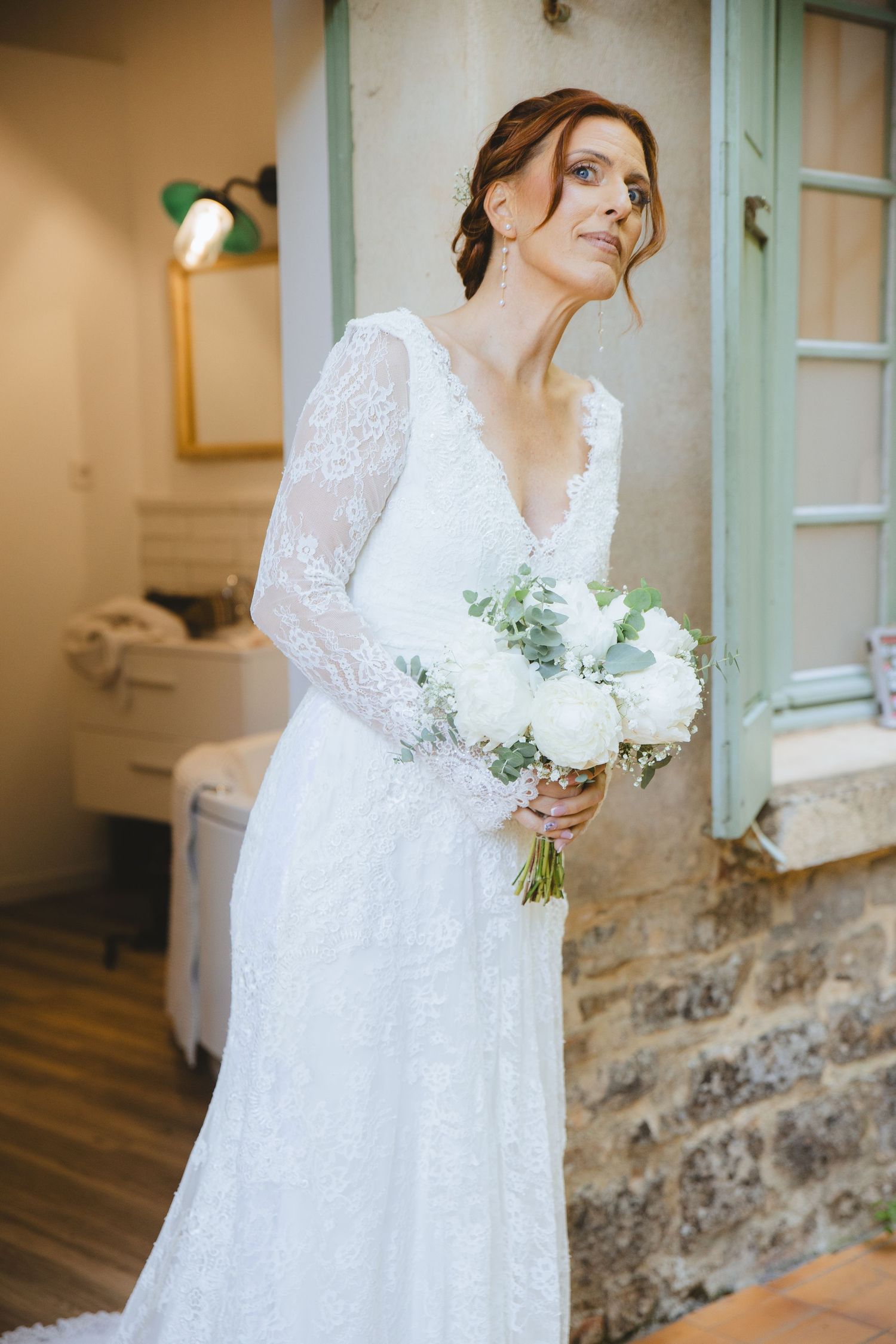 Bride in lace long-sleeve wedding gown holds white floral bouquet, standing near rustic stone wall indoors.