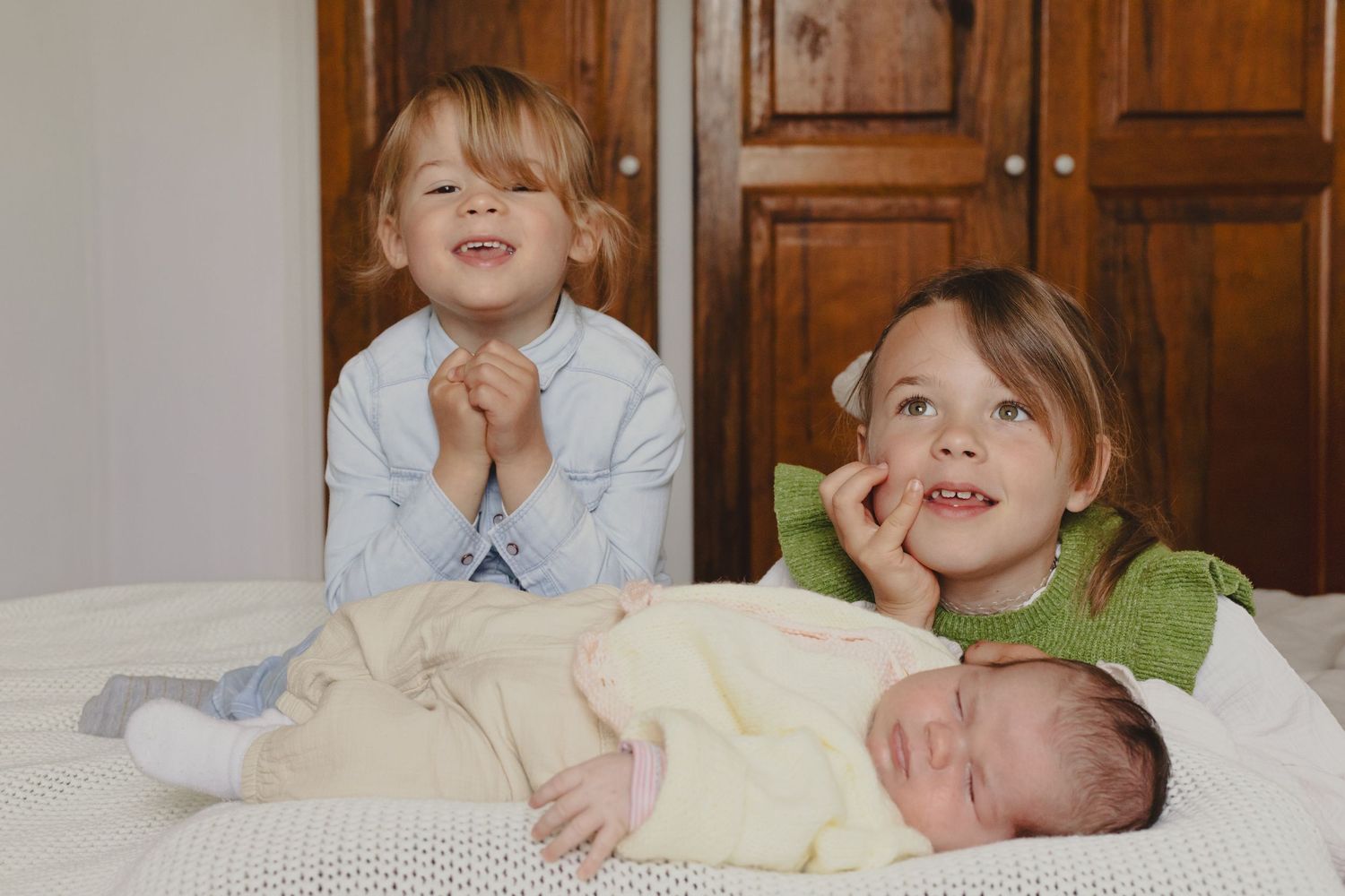 Two young children posing with a sleeping newborn baby on a white bed indoors.