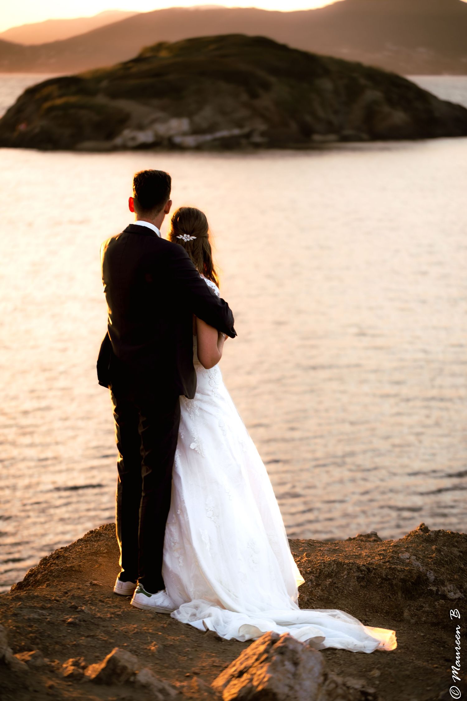 Wedding couple embracing on rocky shore at sunset with island and calm water in background.
