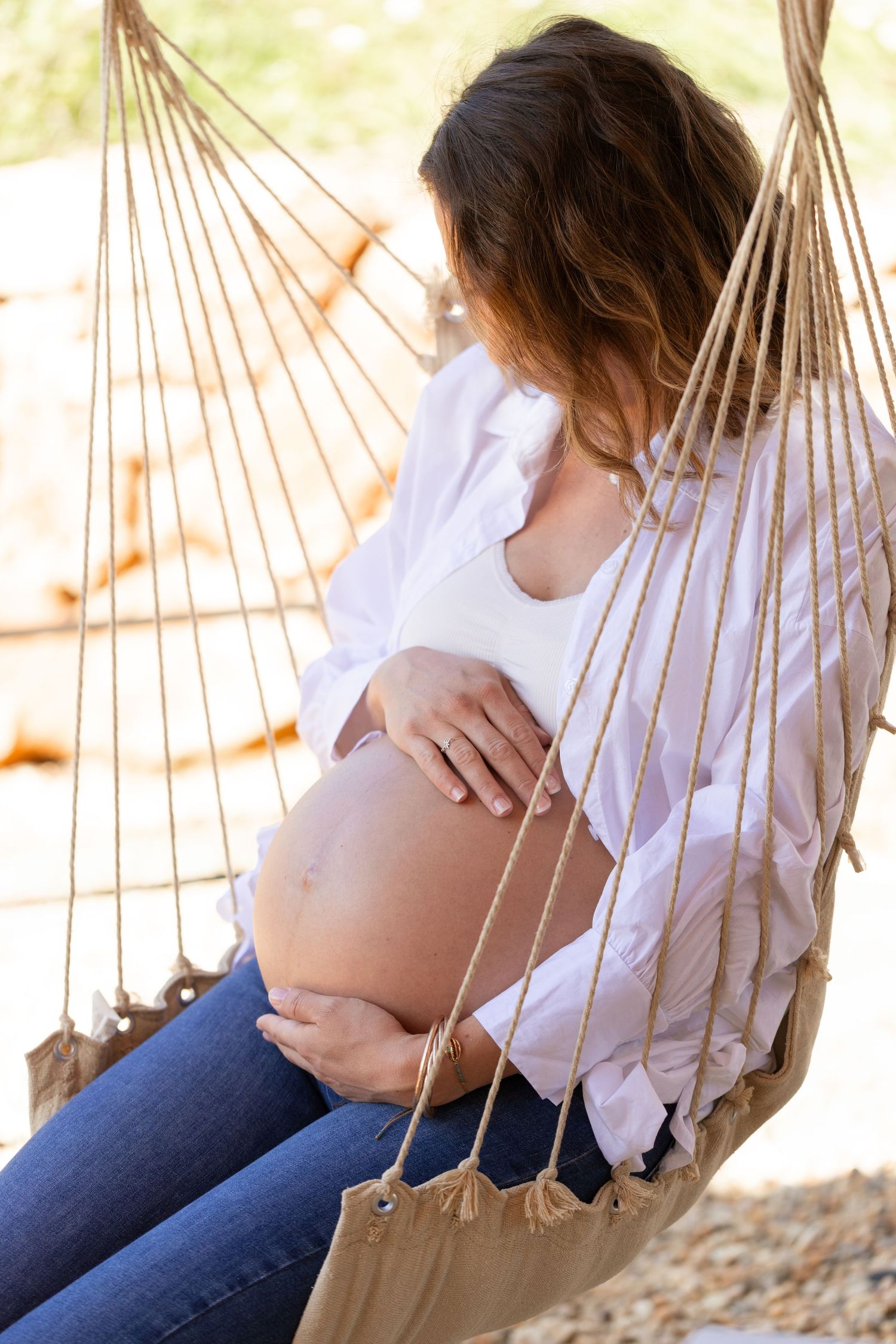 Pregnant woman in white shirt and jeans sitting in hammock outdoors, gently holding her bare belly.