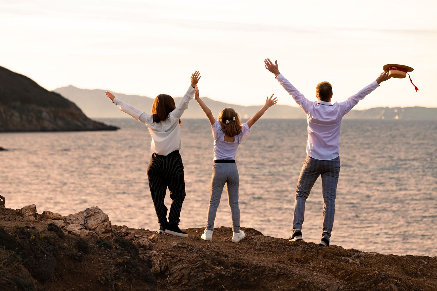 Three people stand on rocky shore with arms raised celebrating at sunset over calm ocean waters.