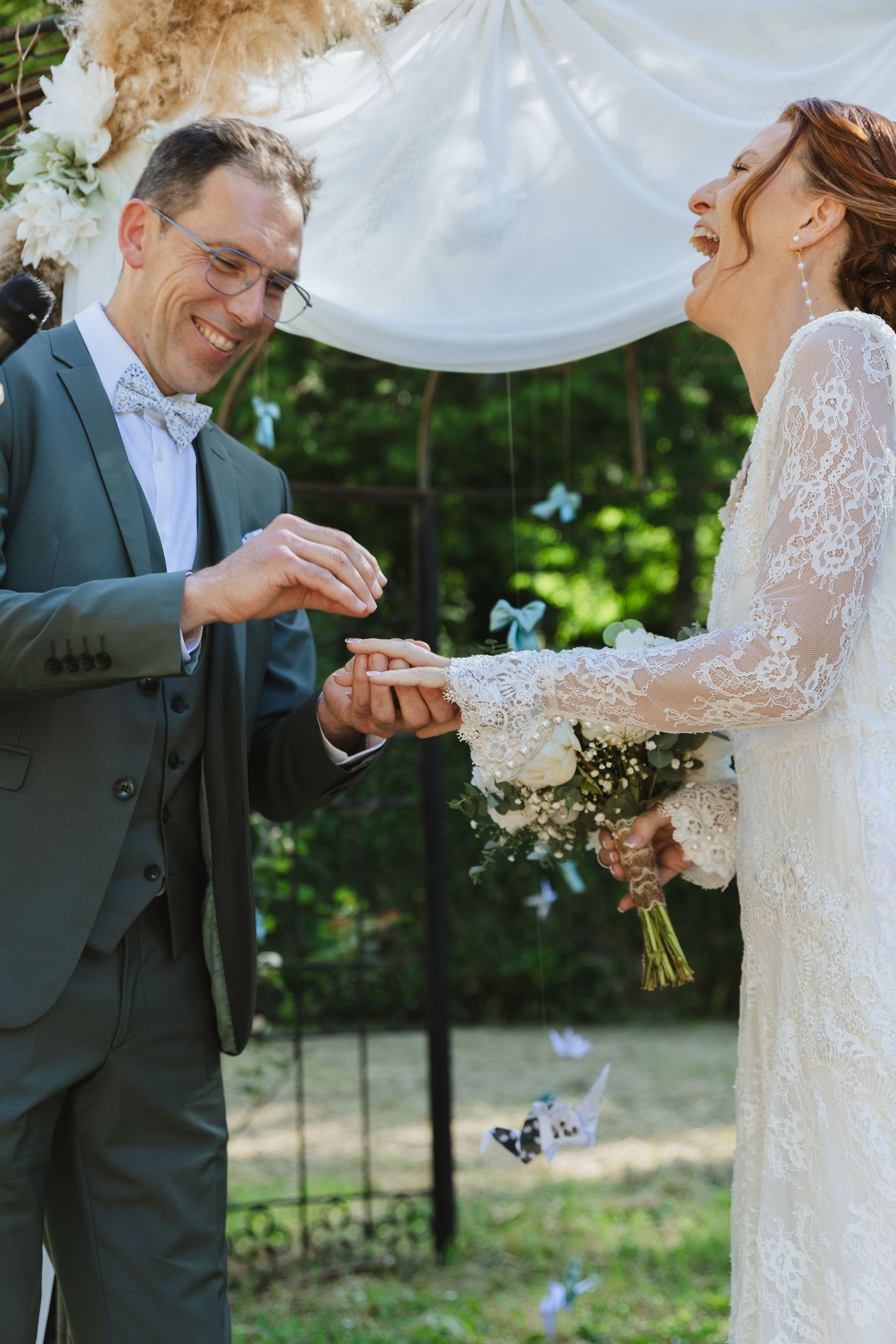 Groom in gray suit places wedding ring on brides finger as she laughs joyfully during outdoor ceremony.