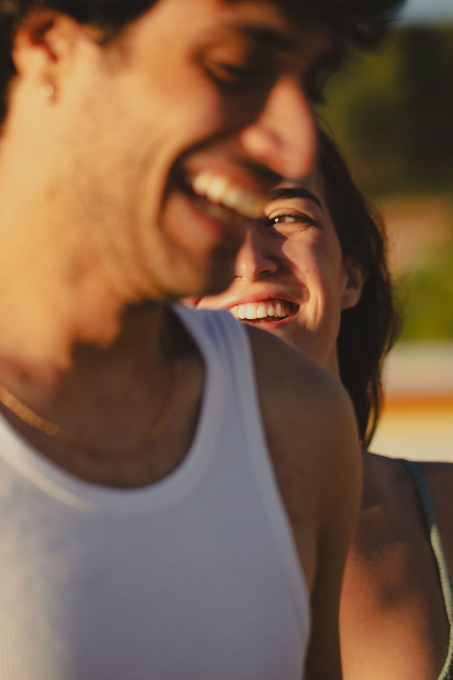 A laughing couple enjoying a sunny day outdoors, with a woman smiling warmly in the background.