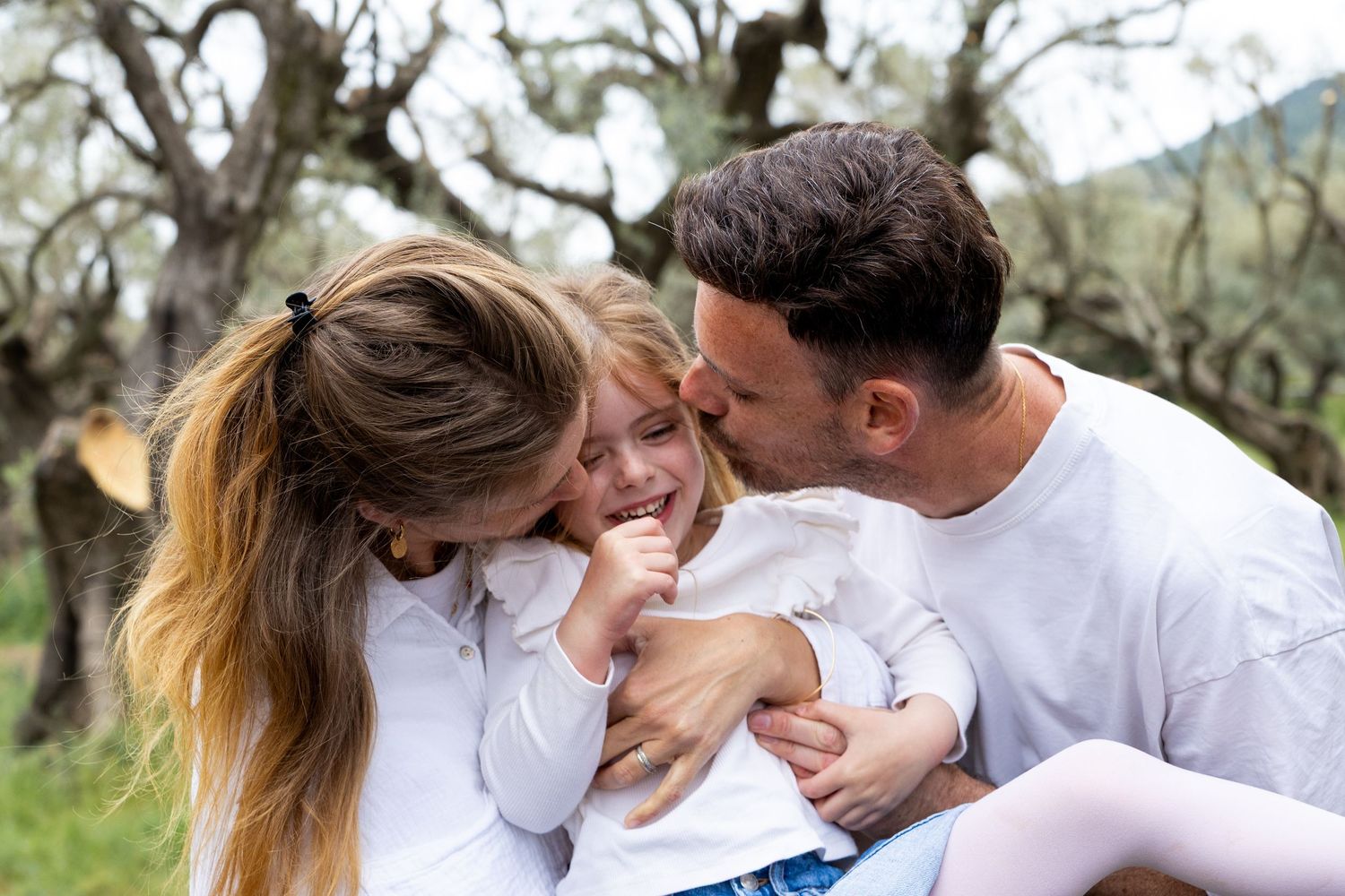 A loving family shares a joyful moment outdoors, with parents kissing their young daughter among trees.