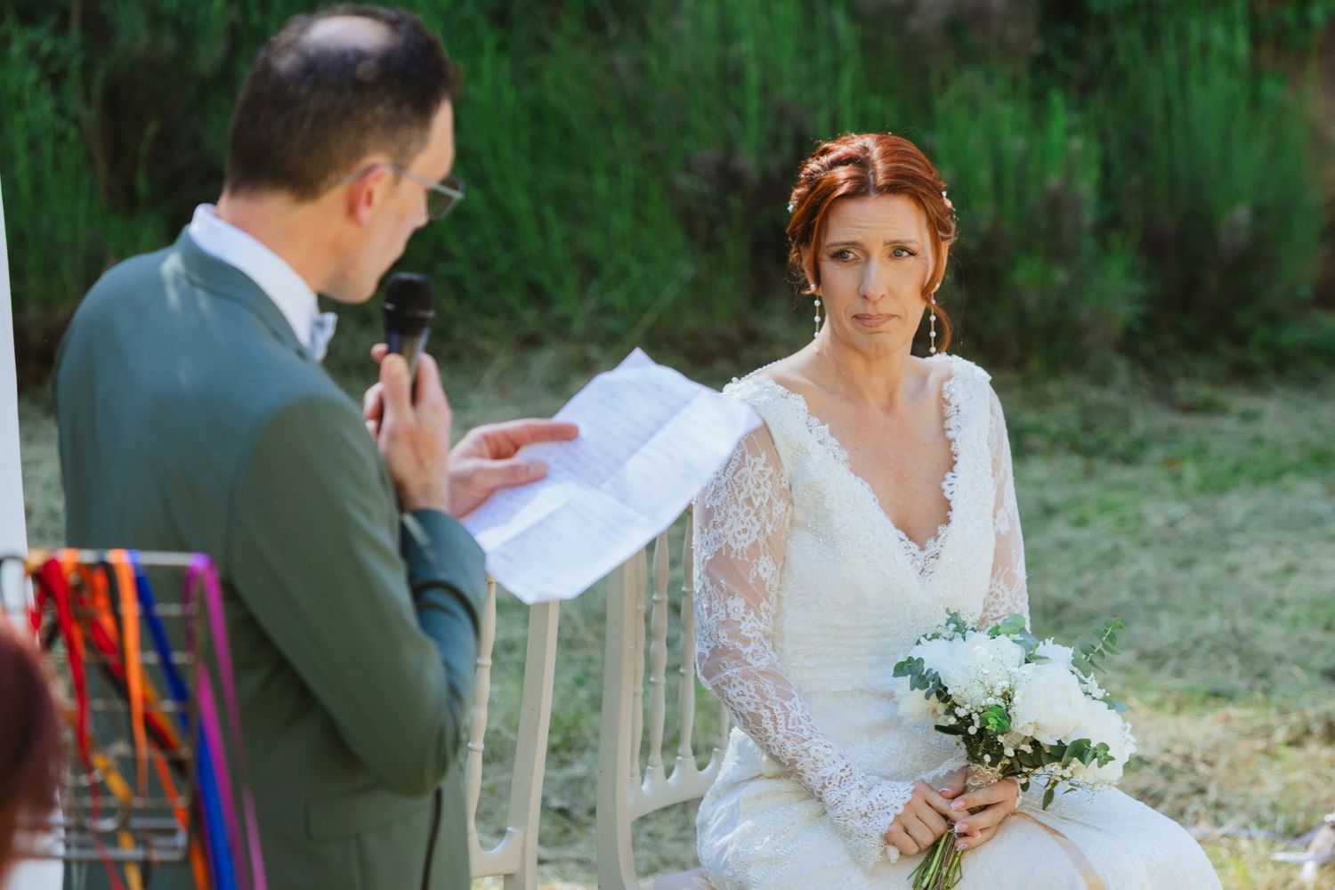 Bride in lace wedding dress holds bouquet while groom reads vows during outdoor wedding ceremony.