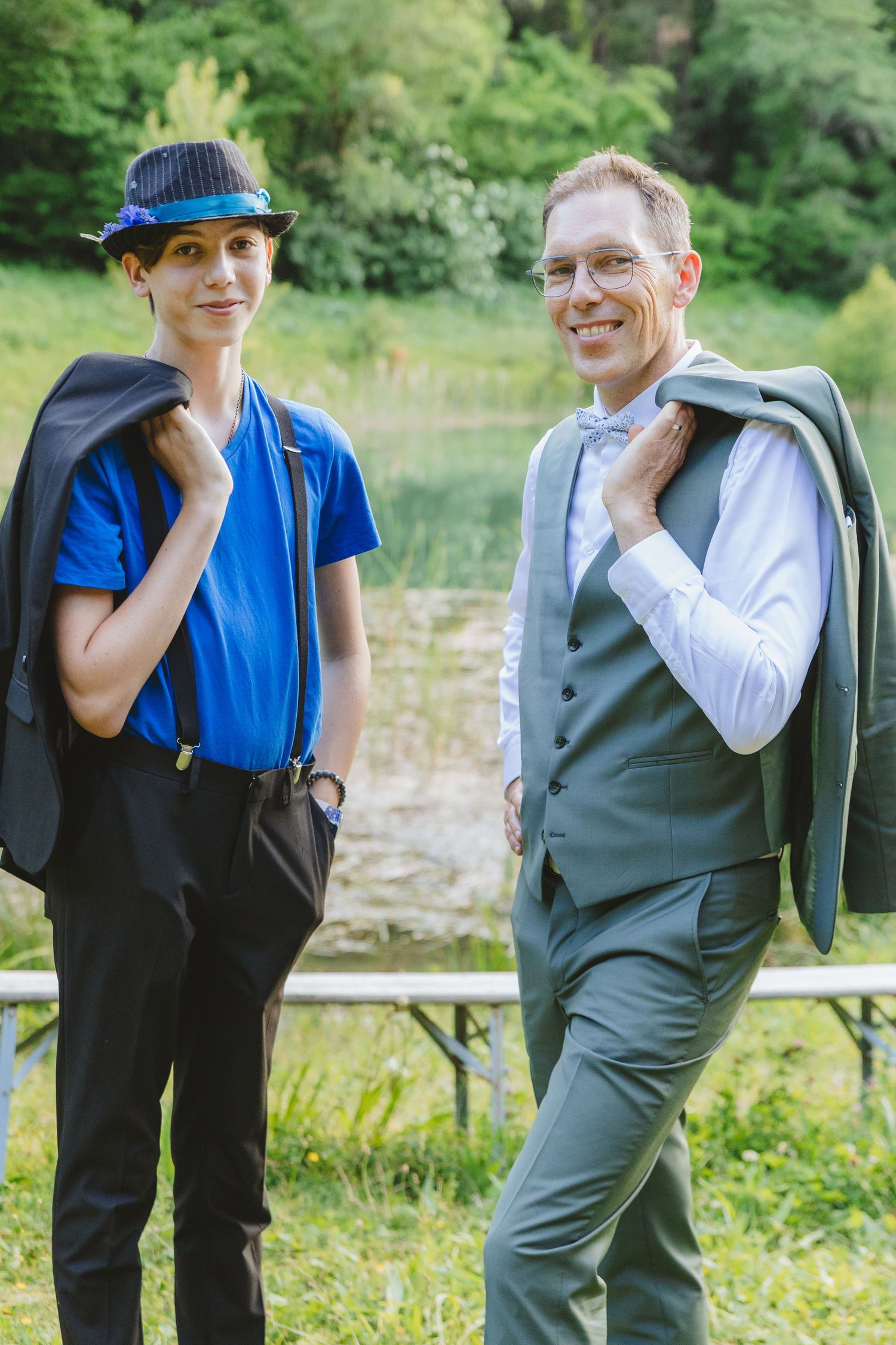 Two people in semi-formal attire holding jackets over shoulders outdoors near a pond with lush greenery.