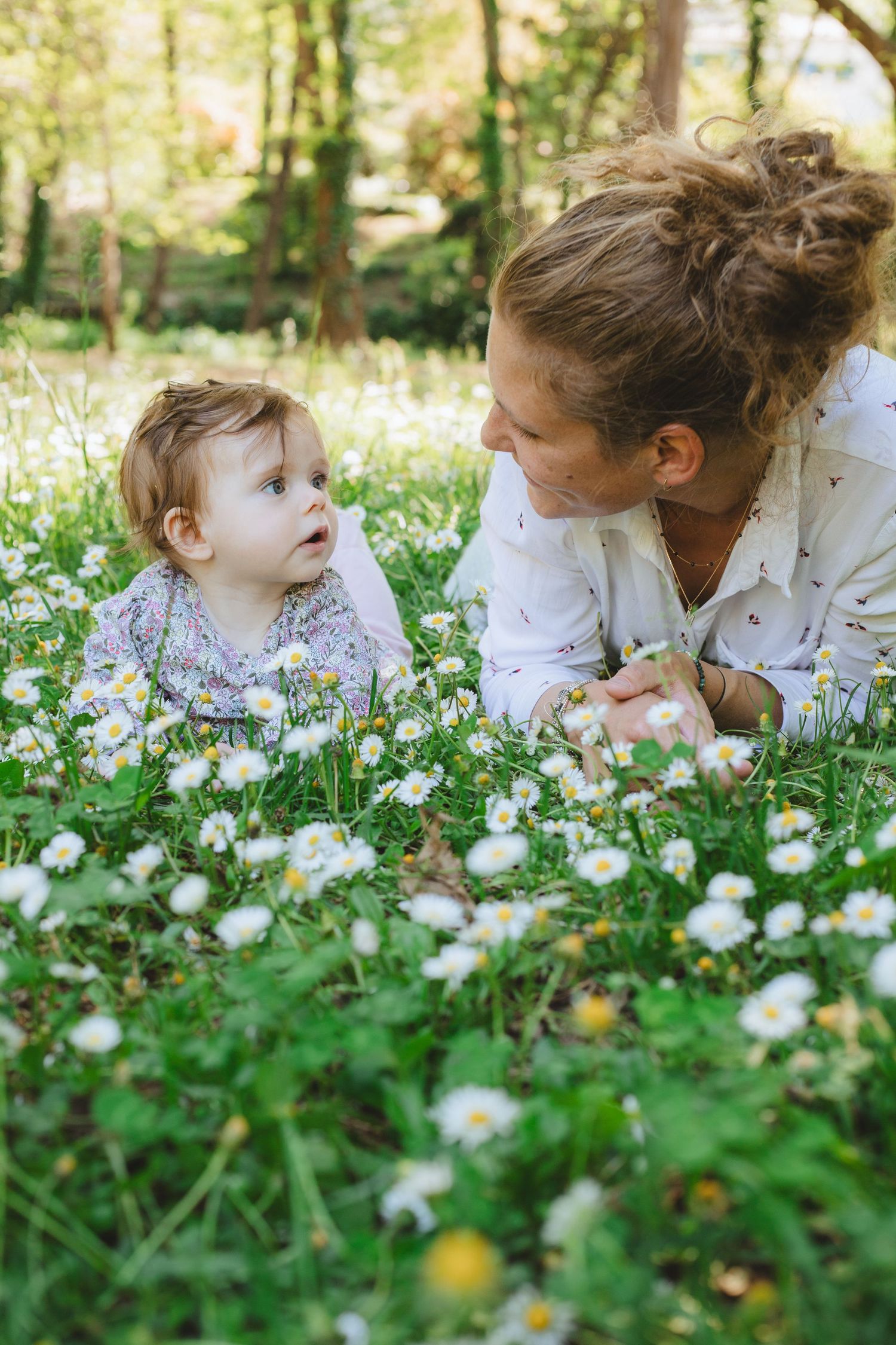 A curly-haired mother lies in a daisy field smiling at her curious baby girl.
