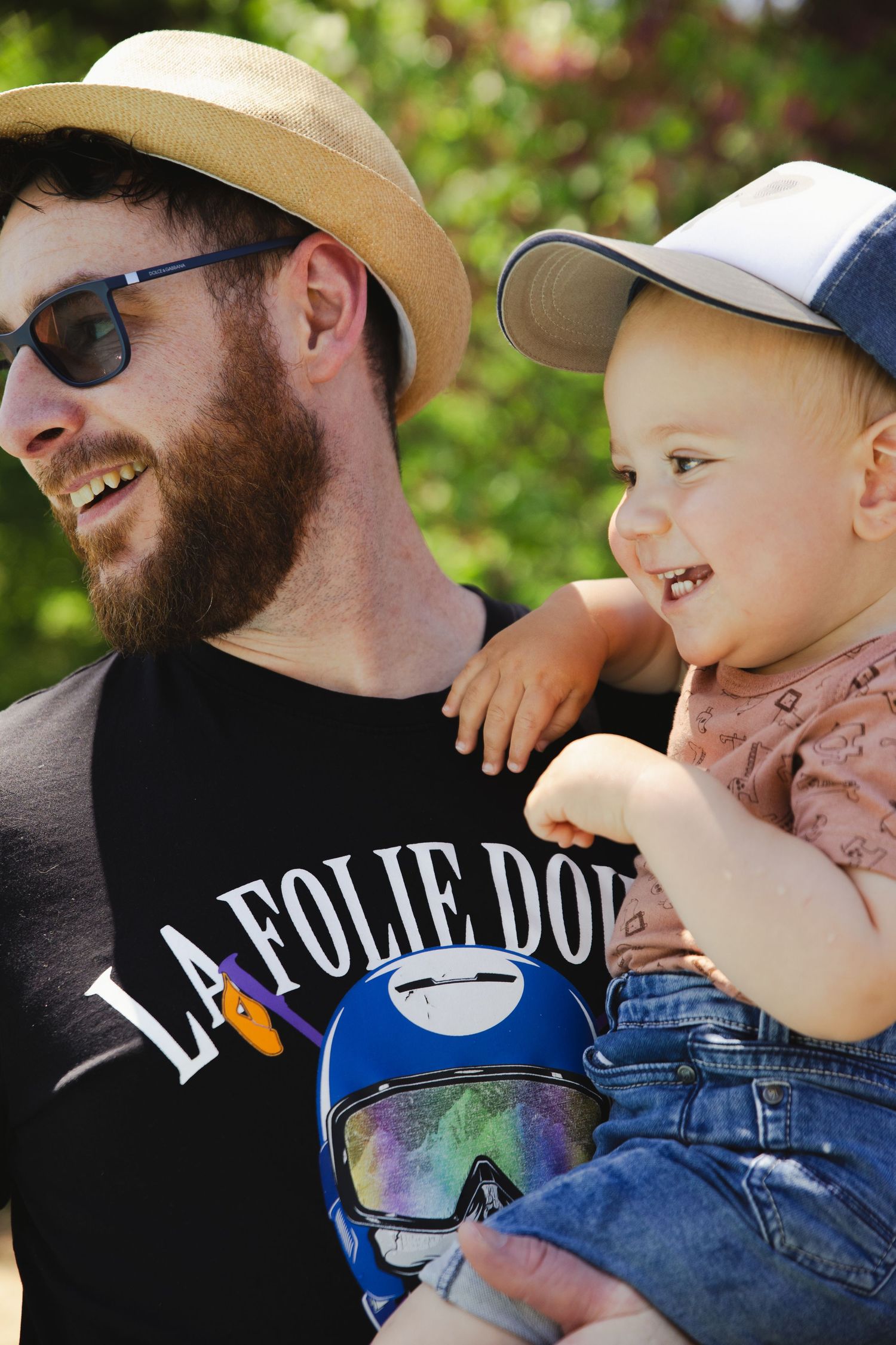 Bearded man in straw hat and sunglasses holding a smiling toddler wearing a cap outdoors.