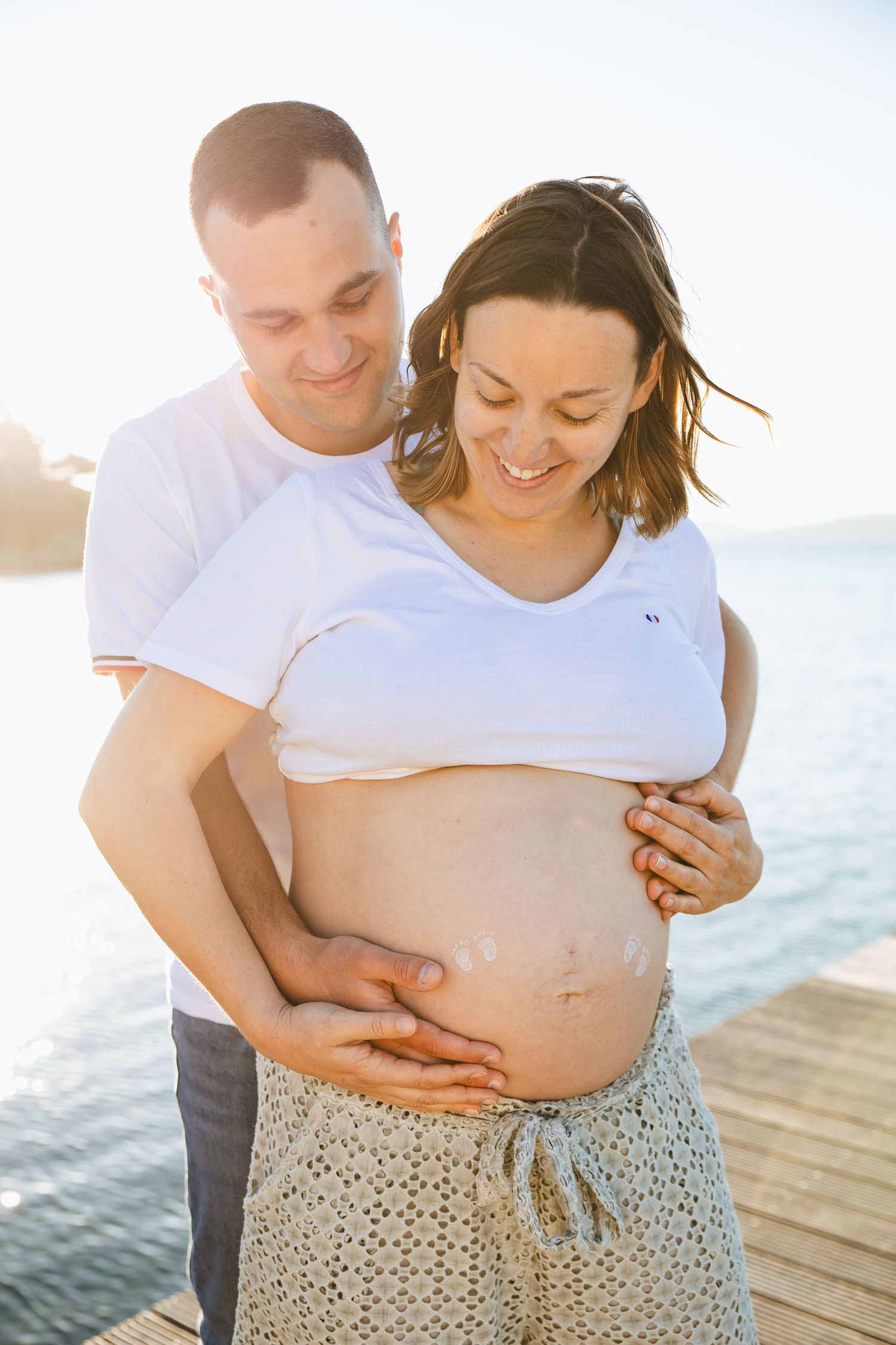 Happy pregnant couple on a sunny pier, man embracing woman's bare baby bump near sparkling water.