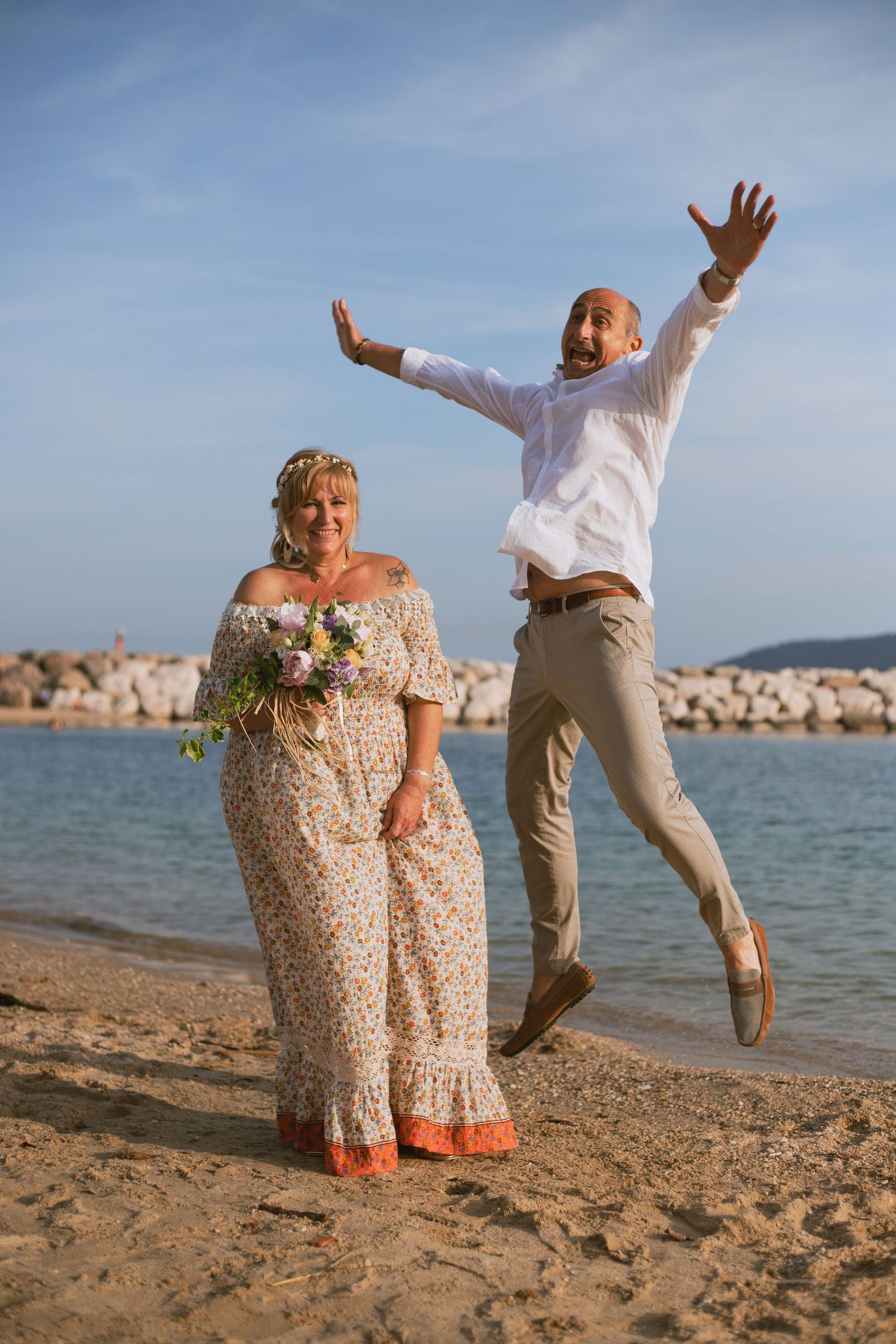 Joyful groom jumping with arms raised beside bride holding bouquet on sandy beach.