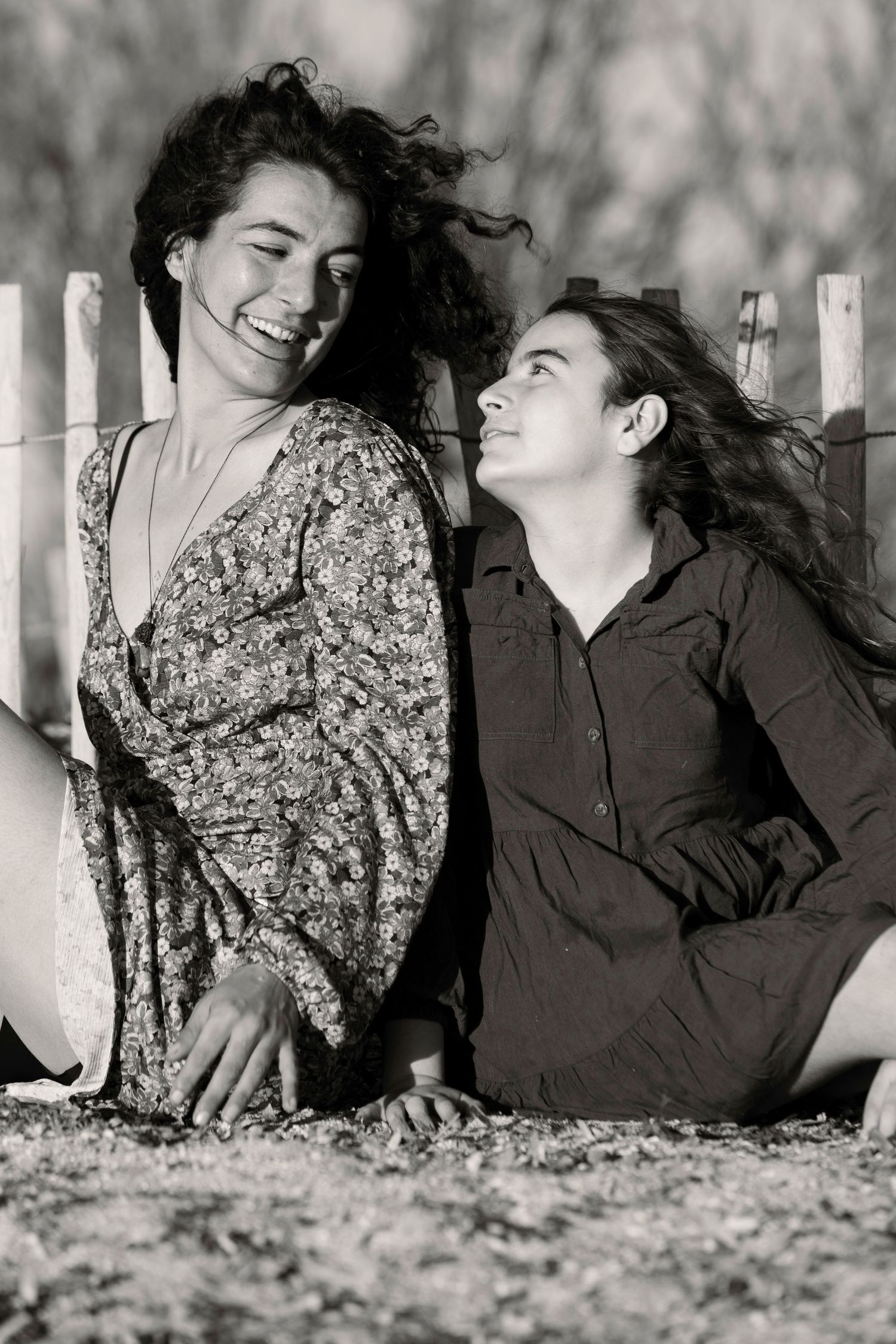 Two young women laugh together leaning against a wooden fence in black and white photo.