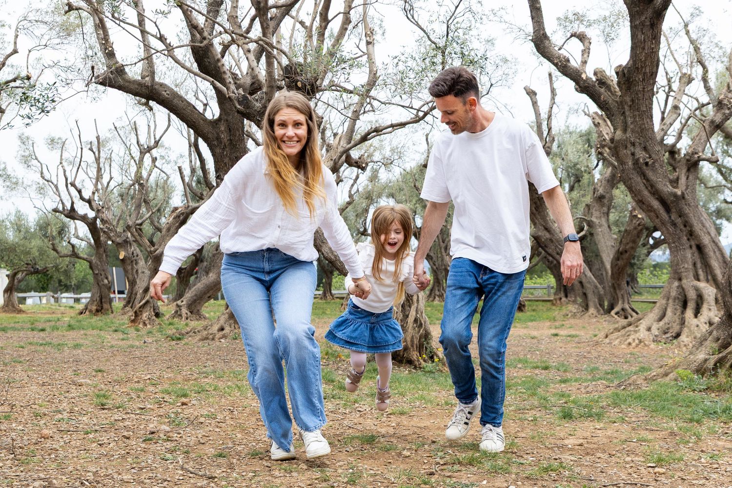 A happy family of three holds hands and walks together through an olive tree park.