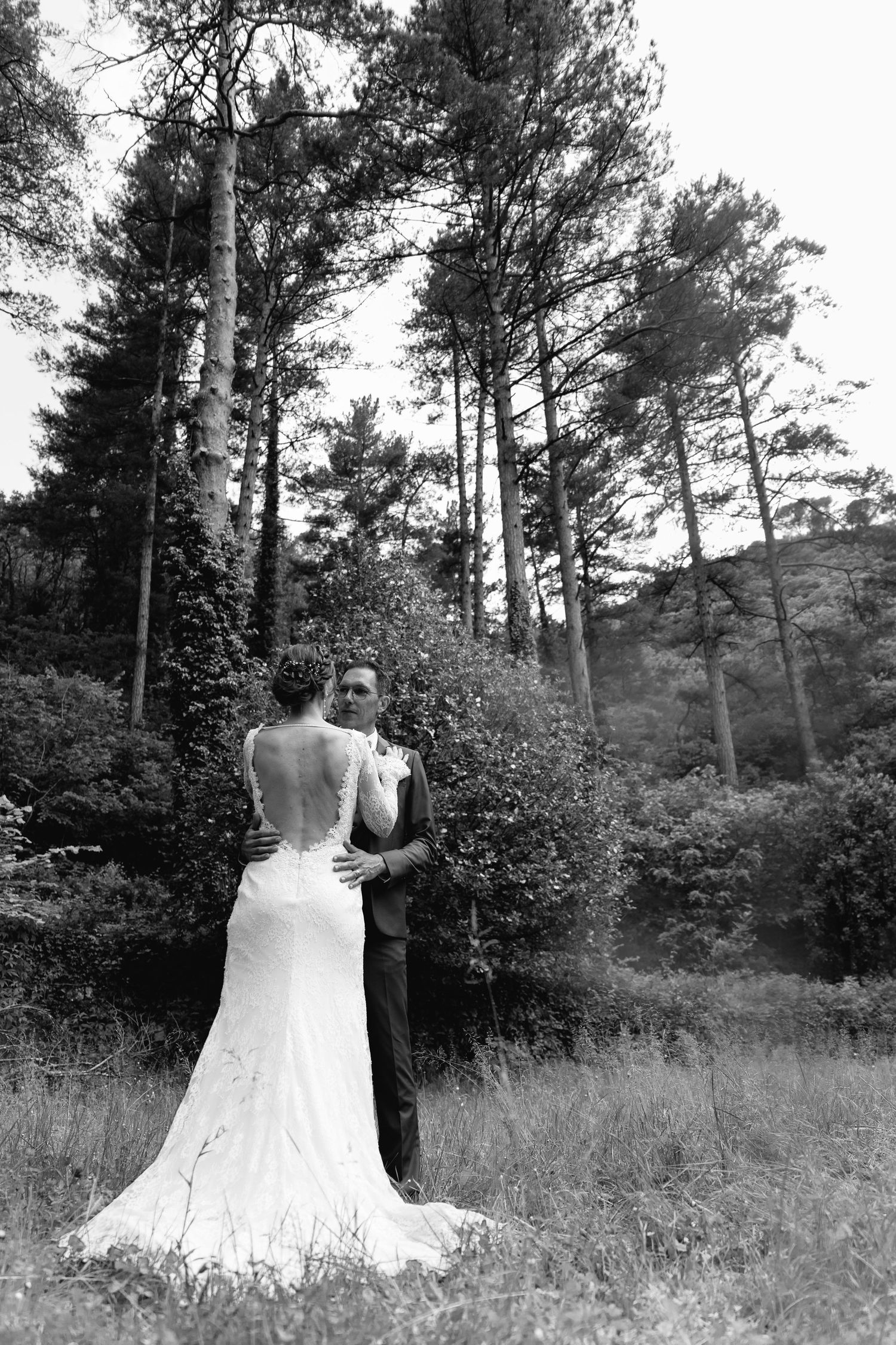 Black and white photo of a bride and groom embracing in a lush forest setting, bride wearing an elegant backless gown.
