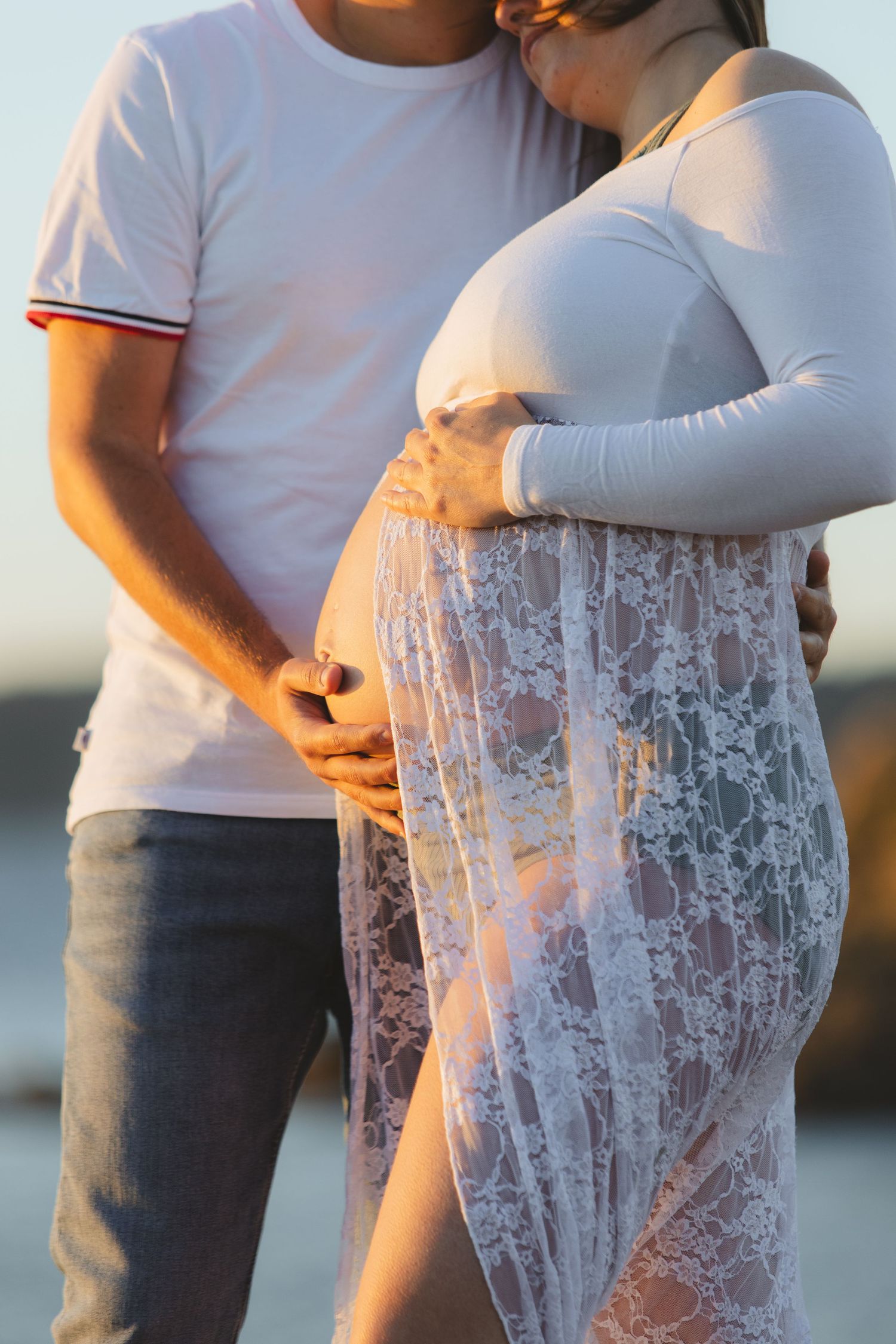 Couple embracing at sunset, man kissing pregnant woman's head while holding her lace-covered baby bump.